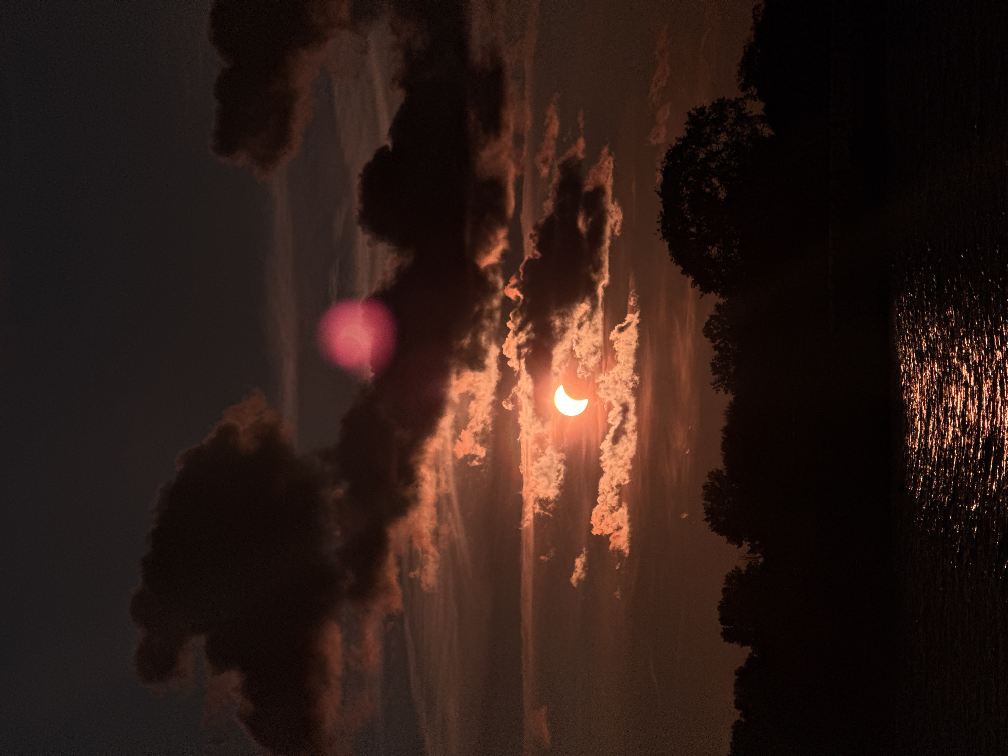 The image captures a partial solar eclipse occurring during sunset over a body of water. The sky is dark with dramatic clouds tinged with orange and red hues from the setting sun. The sun itself is partially obscured by clouds, appearing as a crescent shape. In the foreground, the dark silhouette of a tree line is visible, with the surface of the water reflecting the warm colors of the sky, creating a shimmering effect. There is a noticeable lens flare or atmospheric distortion as a bright pinkish orb in the upper portion of the image. The scene is devoid of any human presence. It is not possible to determine the location in Riga, Latvia from the image provided.