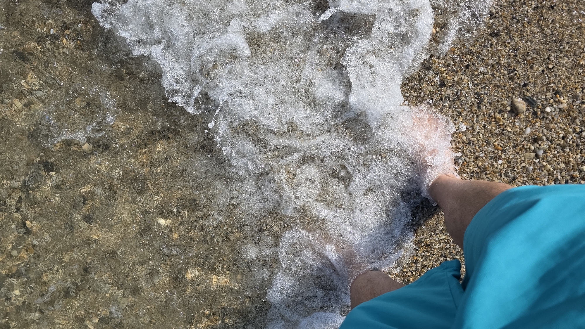 An overhead photograph depicts a person's lower legs and feet partially submerged in the shallow, clear waters of a beach in Asprovalta, Greece. The individual is wearing turquoise swim shorts, visible on the lower-left quadrant of the frame. A white, foamy wave breaks around their feet, creating numerous bubbles and splashes. The seabed, composed of small, varied-colored pebbles and coarse sand, is clearly visible through the transparent water, particularly in the upper portion where the water is less agitated. The shoreline transitions from wet pebbles to drier ones further up the beach. The scene is brightly illuminated by natural daylight.
