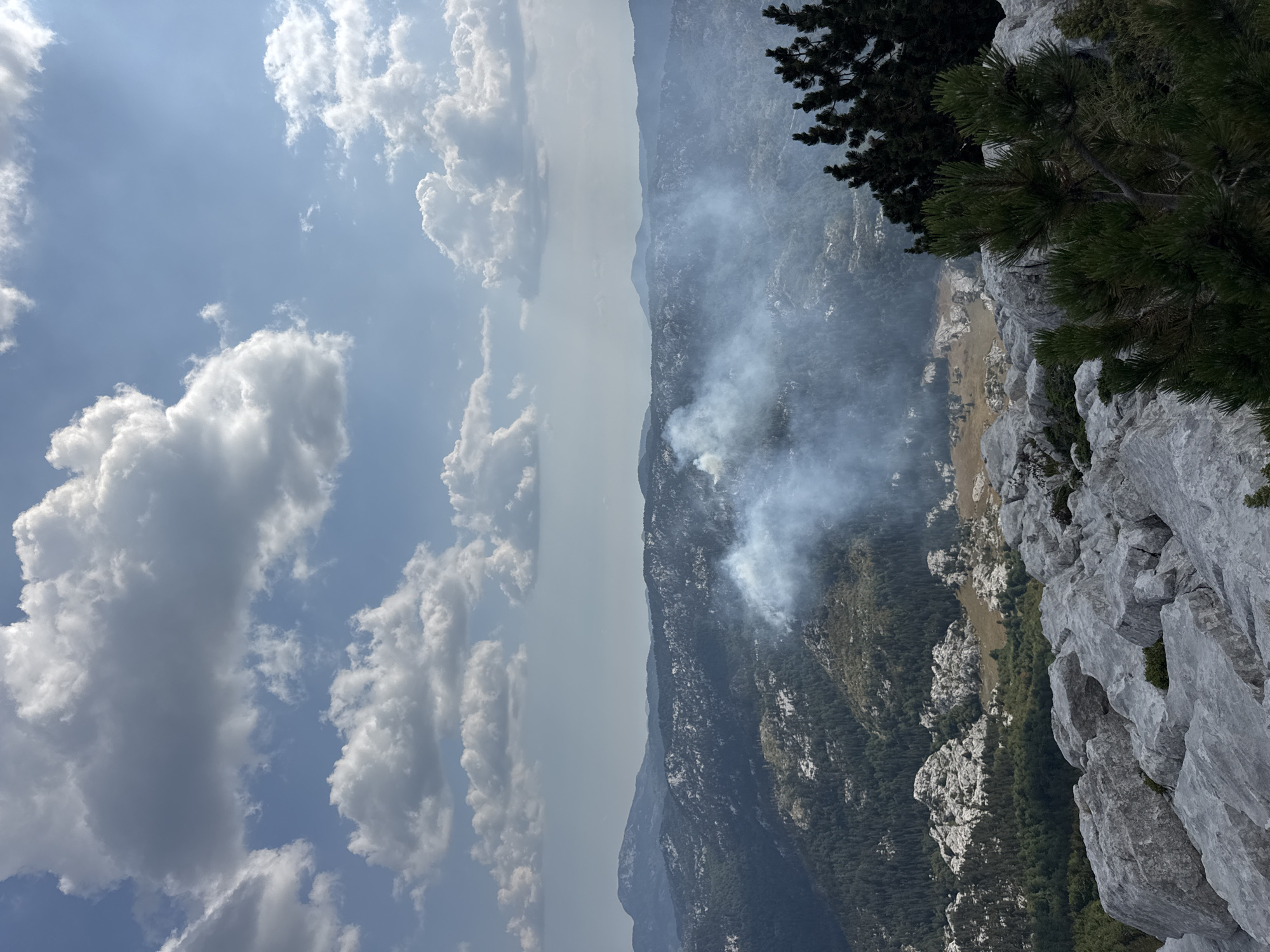 A panoramic, high-angle view captures a hazy mountain landscape under a partly cloudy blue sky. In the mid-ground, a plume of white smoke rises from a densely forested area, suggesting a fire. The foreground consists of rugged, light-grey rock formations and scattered green pine trees. The background reveals rolling, tree-covered mountains that fade into a hazy horizon. The scene is located in the vicinity of Mrtvo Duboko, Montenegro. No people or specific actions are visible.