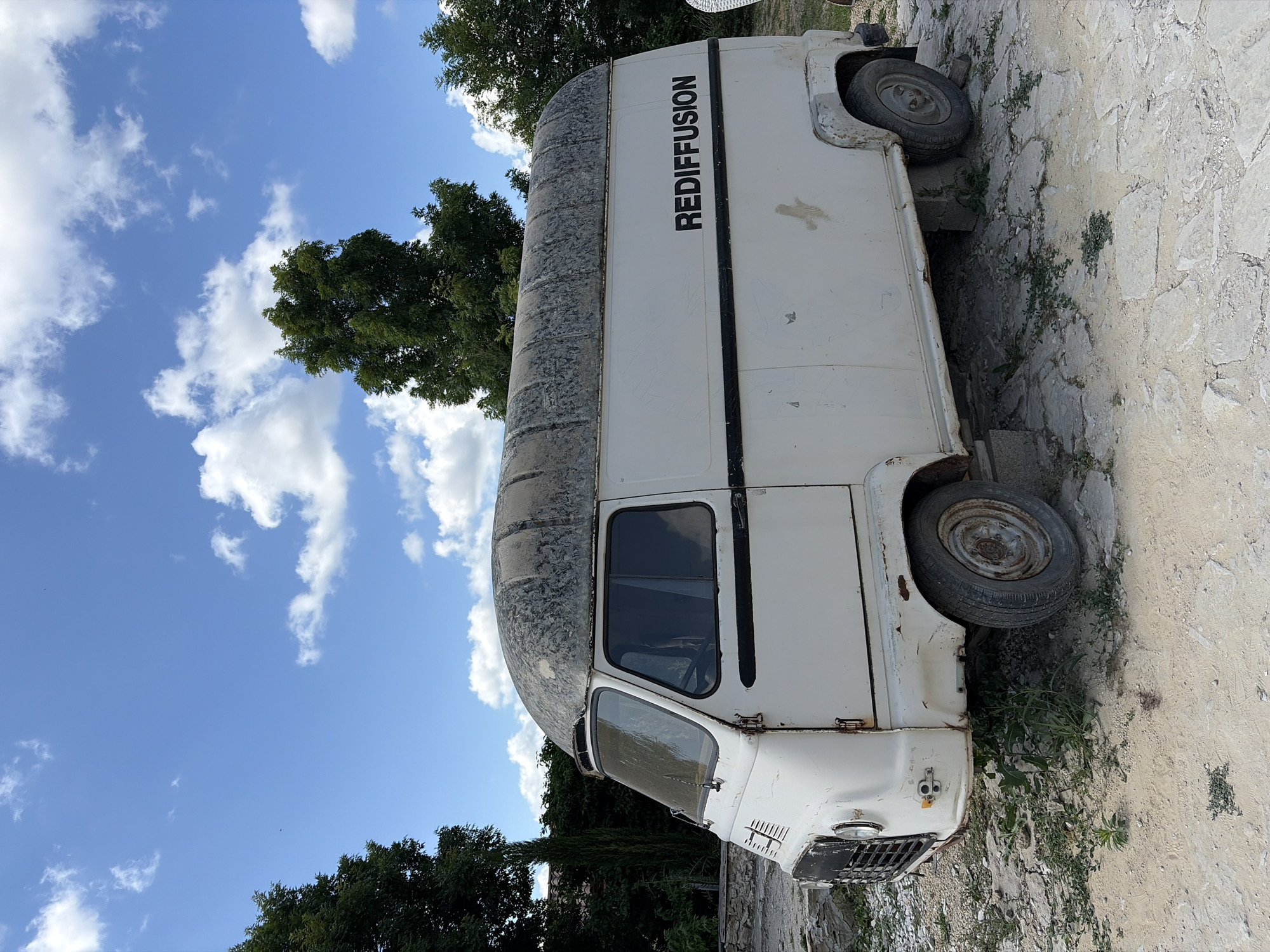 A white, vintage van with the word "REDIFFUSION" in black lettering on its side is parked outdoors. The van is old and appears weathered, with rust visible on some parts. It is situated on a ground covered with sand and stone, with grass growing in patches. In the background, there are trees and a blue sky with white clouds. The image seems to be taken during the daytime. No people are present in the image. The location is identified as Semogorskiy, Russia.