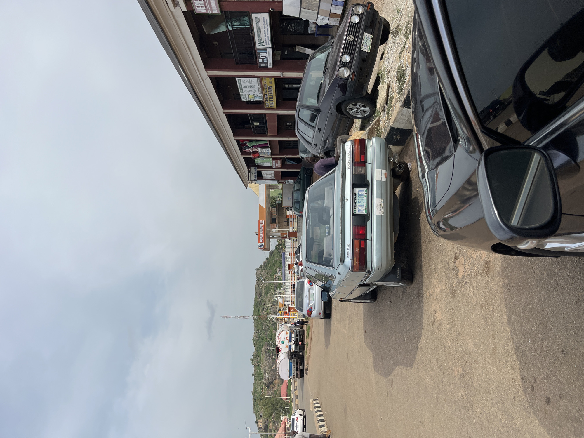 This media file captures a street scene in Gura Top, Nigeria, likely during the daytime with an overcast sky. The foreground is dominated by parked cars. On the right, a dark-colored car's side and side mirror are prominently visible, with its reflection showing other vehicles. To its left, a light-blue Toyota sedan is parked, displaying its rear bumper and license plate which reads "BSA-518EA". The text "TOYOTA" is visible on its tire. Further down the street, another light-blue Toyota sedan is parked, with "TOYOTA" visible on its tire.
Behind the parked cars, a multi-story building with signs is visible. One sign reads "JERE BUILDING", and another, partially obscured, mentions "JDS MULTI-CONCEPTS LTD". Further down, a sign for "TOKO GLOBAL MULT BAGNESS UTI FERTILIZERS SILD HE" is present. A dark-colored Volkswagen Golf is also parked in front of this building, with its license plate reading "JJN-603AG".
In the background, several tanker trucks are lined up, suggesting a logistics or distribution point. The landscape behind the trucks shows a rocky, hilly terrain typical of the region. People are visible in the mid-ground, standing near the vehicles. The overall scene depicts a busy street with parked vehicles, commercial establishments, and transportation infrastructure in a Nigerian setting.