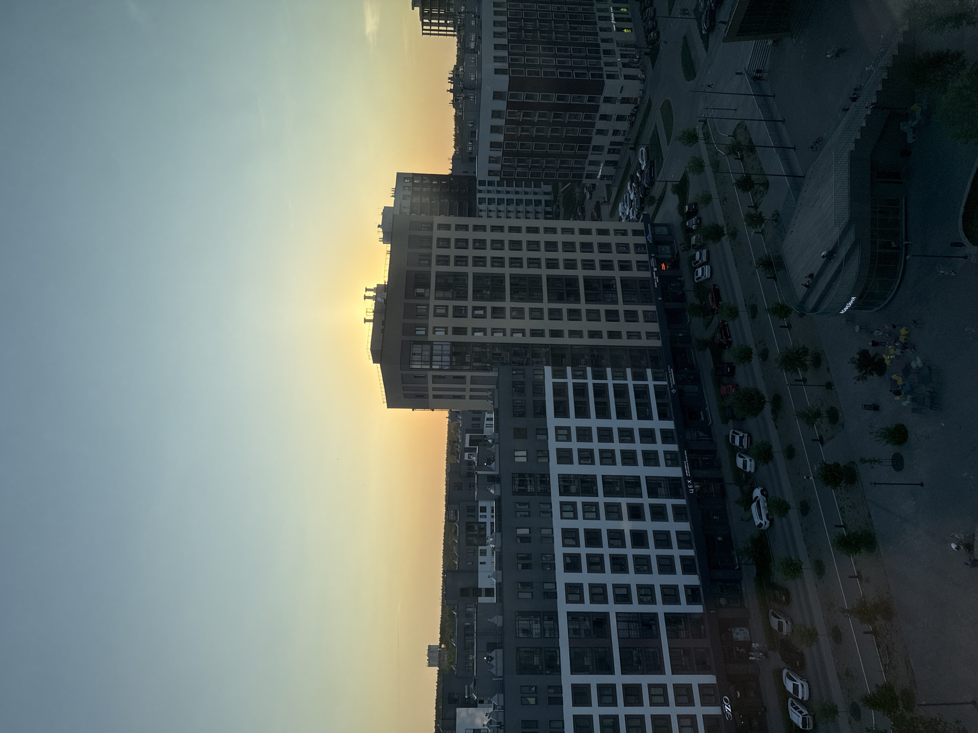 The image presents an elevated view of a city scene, likely captured during dusk or dawn due to the sky gradient from blue to yellow-orange. Modern high-rise buildings dominate the landscape, characterized by their grid-like windows and contemporary architectural styles. A street runs parallel to the buildings, lined with trees and parked cars. In the foreground, there's a public space with steps and seating areas, where some people are gathered. The scene appears to be in Izhevsk, Russia, based on the "MANHATTAN" signage.