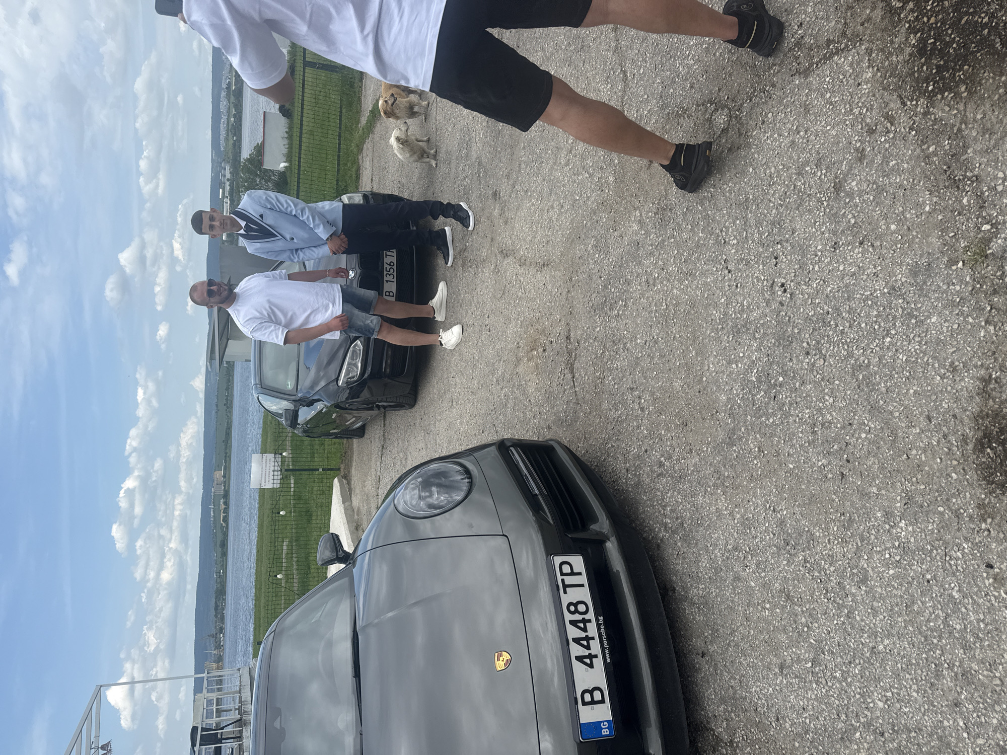 The image shows a group of people and several cars on a paved surface, likely a parking area. The cars include a dark-colored Porsche with a Bulgarian license plate (B 4448 TP) and another car (B 1356 TT). Two men stand near one of the cars, one wearing a white shirt and shorts and the other in a light blue suit jacket. A third person in a white shirt and black shorts is partially visible in the upper portion of the image. In the background, there is a green fence and a body of water, which is likely the Danube River, given the location is Konstantinovo, Bulgaria, according to the prompt. Two dogs are visible in the background.