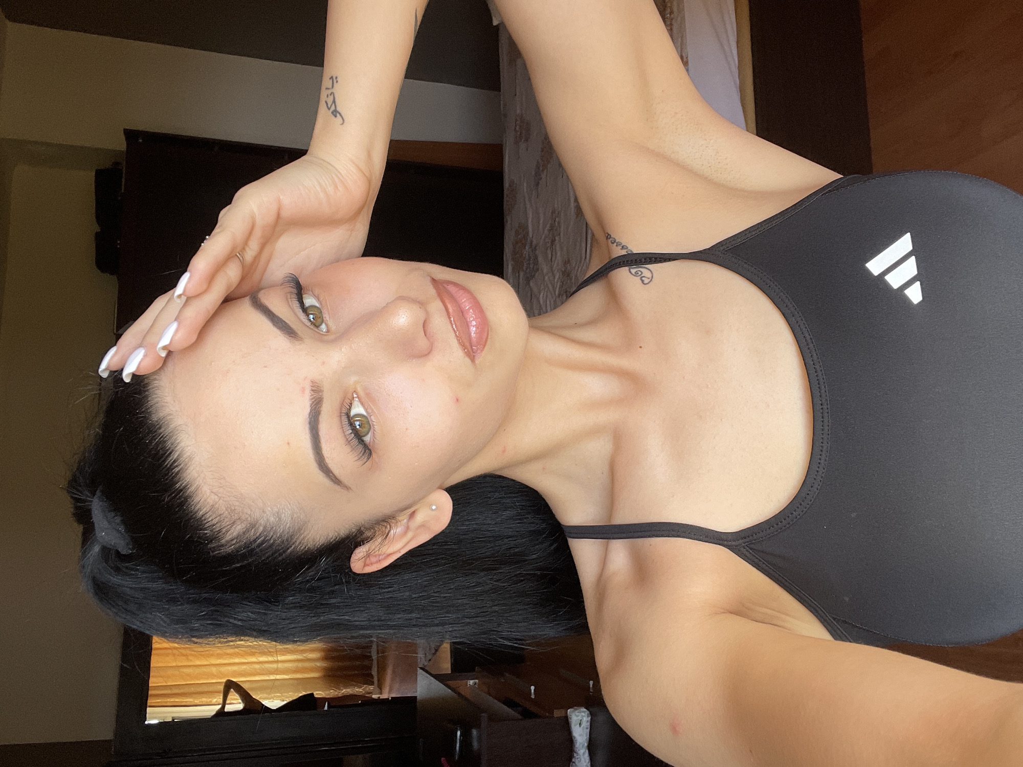 A young woman with black hair pulled into a high ponytail is looking at the camera. She is wearing a black sports bra with a white Adidas logo. Her right hand is resting on her forehead, and she has long fingernails. There are two tattoos visible: one on her left wrist with Arabic script, and another on her right shoulder blade with a heart and vines. She has light-colored eyes and full lips. The background appears to be a bedroom, with a bed and a dresser visible. Curtains hang in the background, and a doorway is visible. The lighting suggests it is daytime. The image does not contain any information indicating its location in Stara Zagora, Bulgaria.