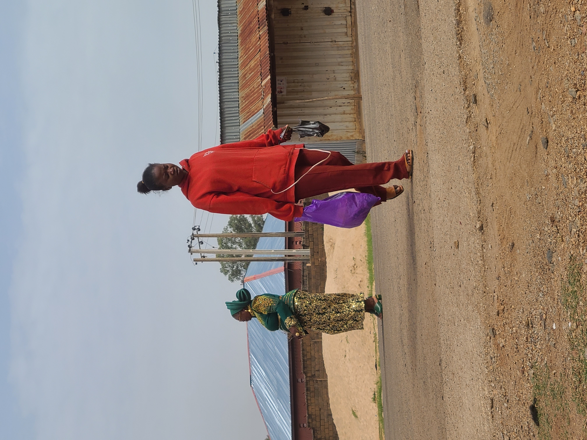 Two women are walking on a paved road with unpaved shoulders in Gura Top, Nigeria. The woman closest to the camera is wearing a green and gold traditional dress. The other woman is wearing a red tracksuit and sandals. She carries a purple bag in one hand and a dark object in the other. Buildings with corrugated metal roofs line one side of the road. Utility poles are visible in the background. The sky is overcast. The ground is mostly dirt and gravel.