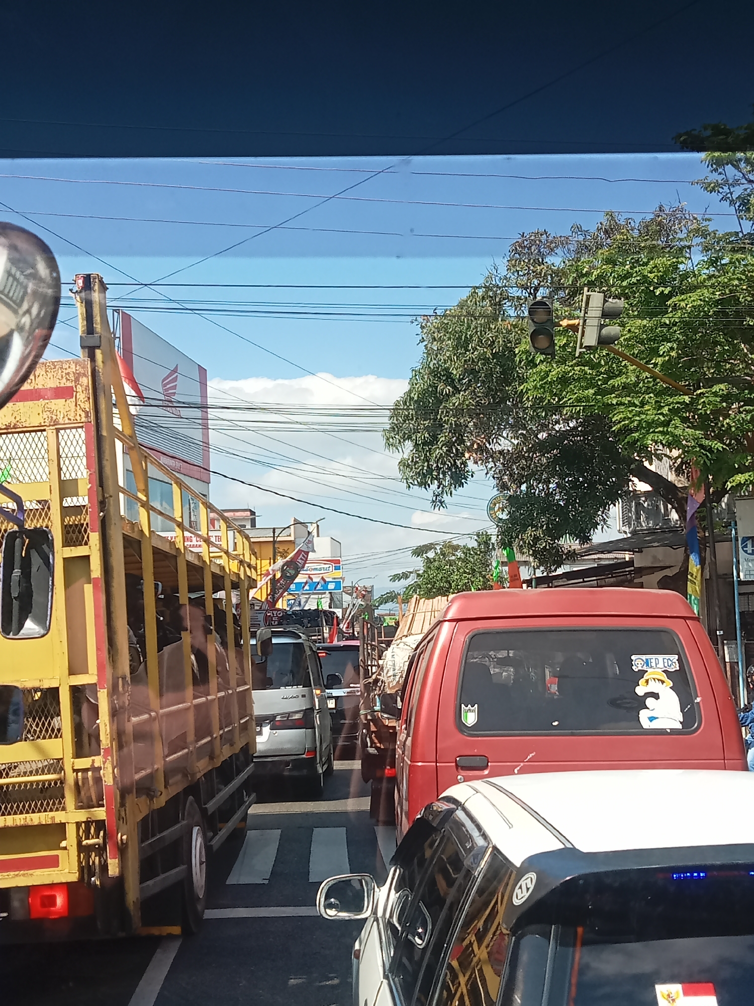 The image captures a view of traffic congestion on a sunny day, likely in Wonosobo, Indonesia, given the Indonesian flag visible on a vehicle. In the foreground, a yellow truck is prominently positioned on the left, its open cargo bed filled with people. To the right of the truck, a red van is visible, displaying stickers on its rear window, including one with a cartoon character that appears to be from "One Piece." Several other vehicles, including a silver minivan and what seems to be a white car, are also present in the traffic jam. Overhead, traffic lights are visible, indicating an intersection. The environment suggests a typical urban or semi-urban street scene with buildings and trees lining the road, and a clear blue sky with some clouds. Power lines are strung across the sky, a common sight in Indonesian cities. The overall impression is one of a slow-moving or stopped traffic situation.