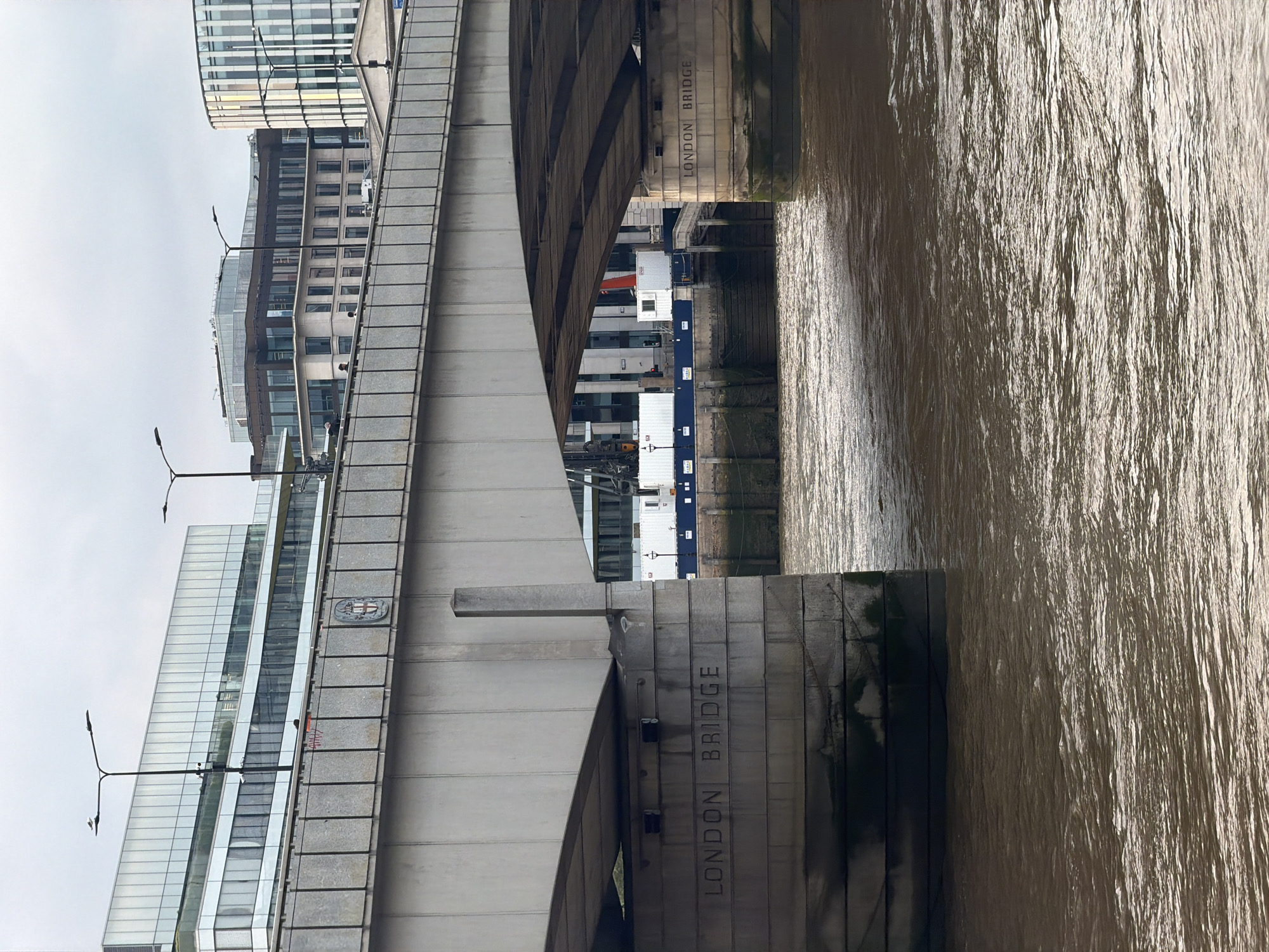 The image shows a view of the London Bridge, situated in London, United Kingdom. The perspective is looking up at a section of the bridge, with the Thames River visible on the right side of the frame. The bridge's concrete structure features the inscription "LONDON BRIDGE" vertically. The river's surface is rippled and reflects the ambient light. On the opposite bank, glimpses of buildings and what appear to be shipping containers can be seen. Street lamps are also visible along the bridge structure. The sky is overcast.
