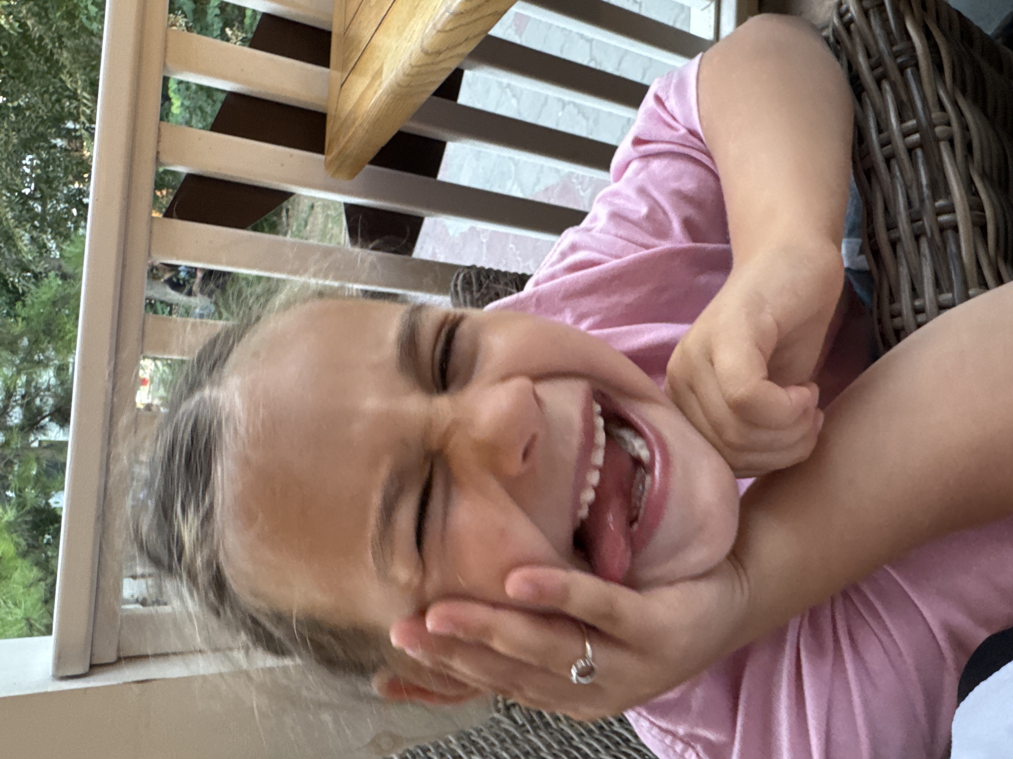 A young girl with blonde hair pulled back is shown in a close-up, outdoor portrait. She is winking, with her mouth open in a wide smile and her tongue sticking out, and she is holding her left cheek with her left hand. She is wearing a pink t-shirt and a silver ring on her left ring finger. The background shows a wooden railing and greenery, suggesting a location such as a porch or balcony. The image appears to have been taken during daylight.