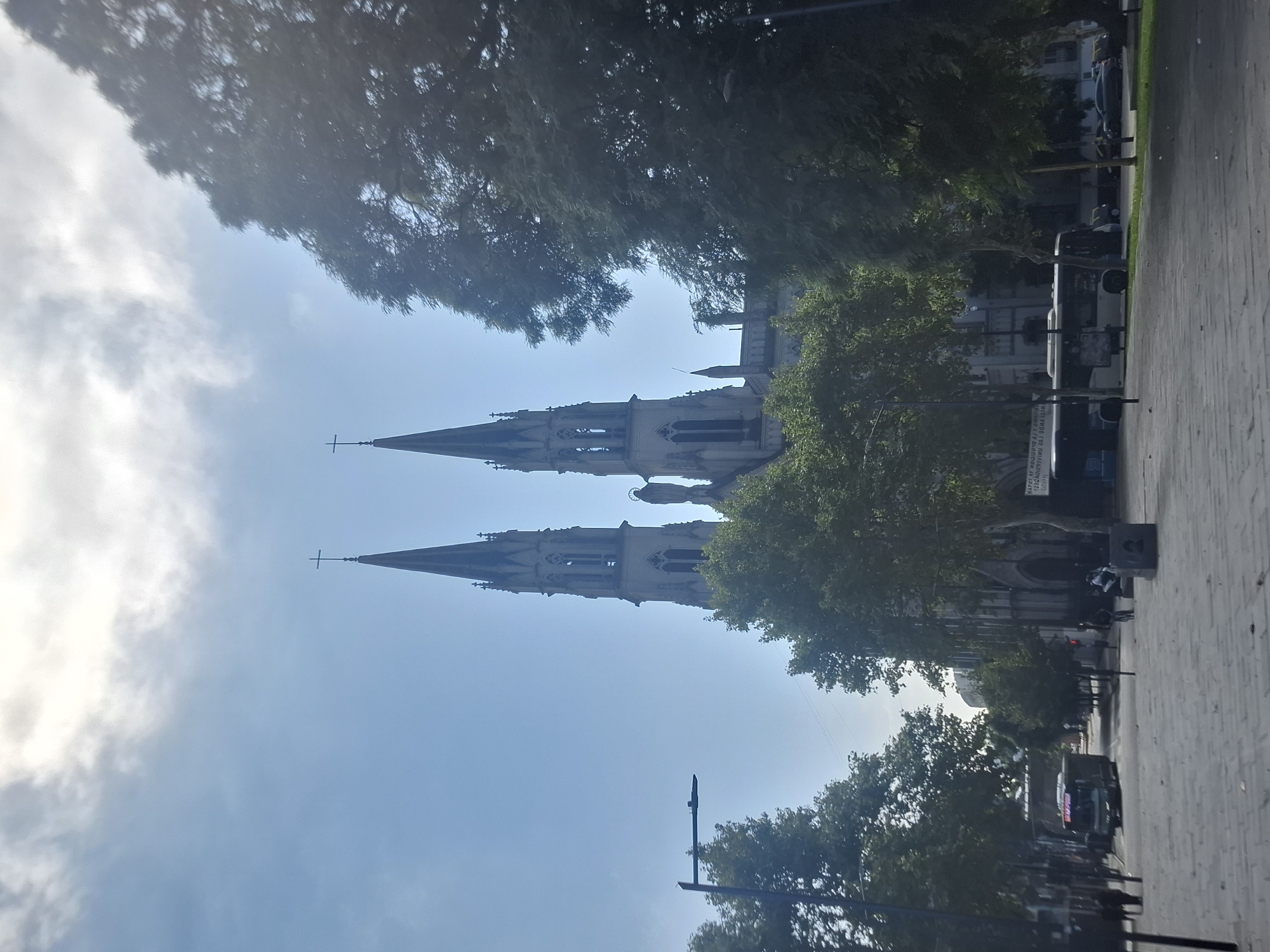 The image captures the Metropolitan Cathedral of Buenos Aires, Argentina, from a slightly low angle on a sunny day. The cathedral's spires dominate the frame, reaching towards the blue sky. Trees partially obscure the view of the building and frame the shot. In the foreground, a pedestrian area with paving stones is visible. Buses and other vehicles are visible in the distance on a street adjacent to the pedestrian zone. A few people can be seen walking along the street.