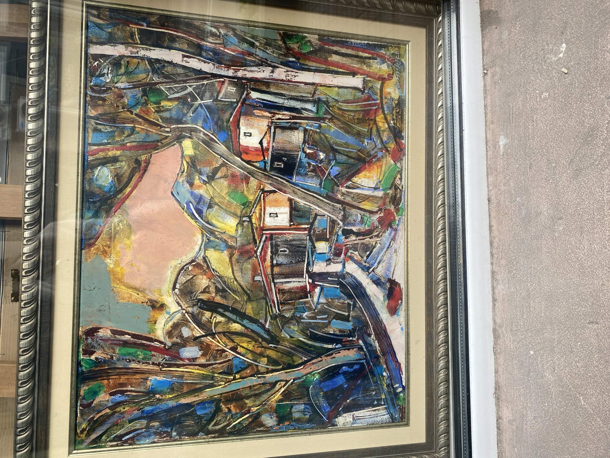 The image shows a framed, abstract oil painting, possibly hanging in a shop window in Varna, Bulgaria. The painting is dominated by a swirling array of colors, including blues, browns, greens, yellows, reds, and a prominent pink or peach area on the left side. The composition features what appear to be stylized buildings or structures, connected by a winding path or road. The painting's style suggests a modern or contemporary approach, with heavy brushstrokes and an emphasis on color and texture rather than realistic representation.
The frame surrounding the painting is ornate and multi-layered, with a silver or metallic finish. The painting is hanging, or placed, behind a window. The background consists of parts of other buildings (window on the left) and the wall on the right, with a grainy, textured surface.