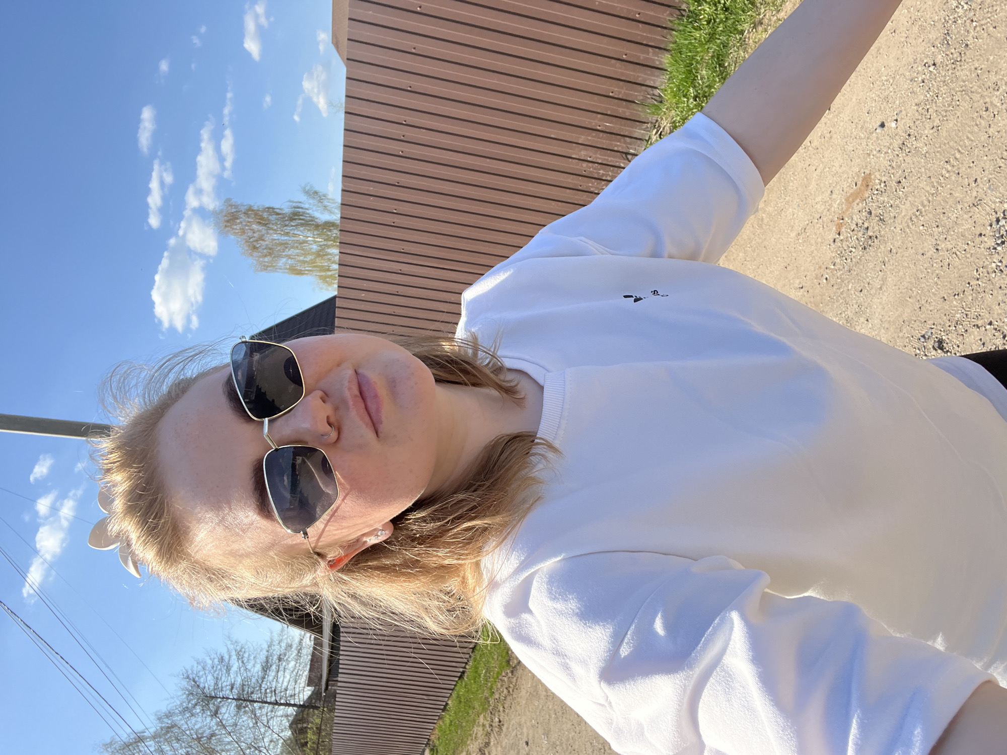 A young woman wearing sunglasses and a white sweatshirt is standing on a dirt road in Chaikovsky, Russia. A brown fence and a blue sky with clouds are visible in the background.