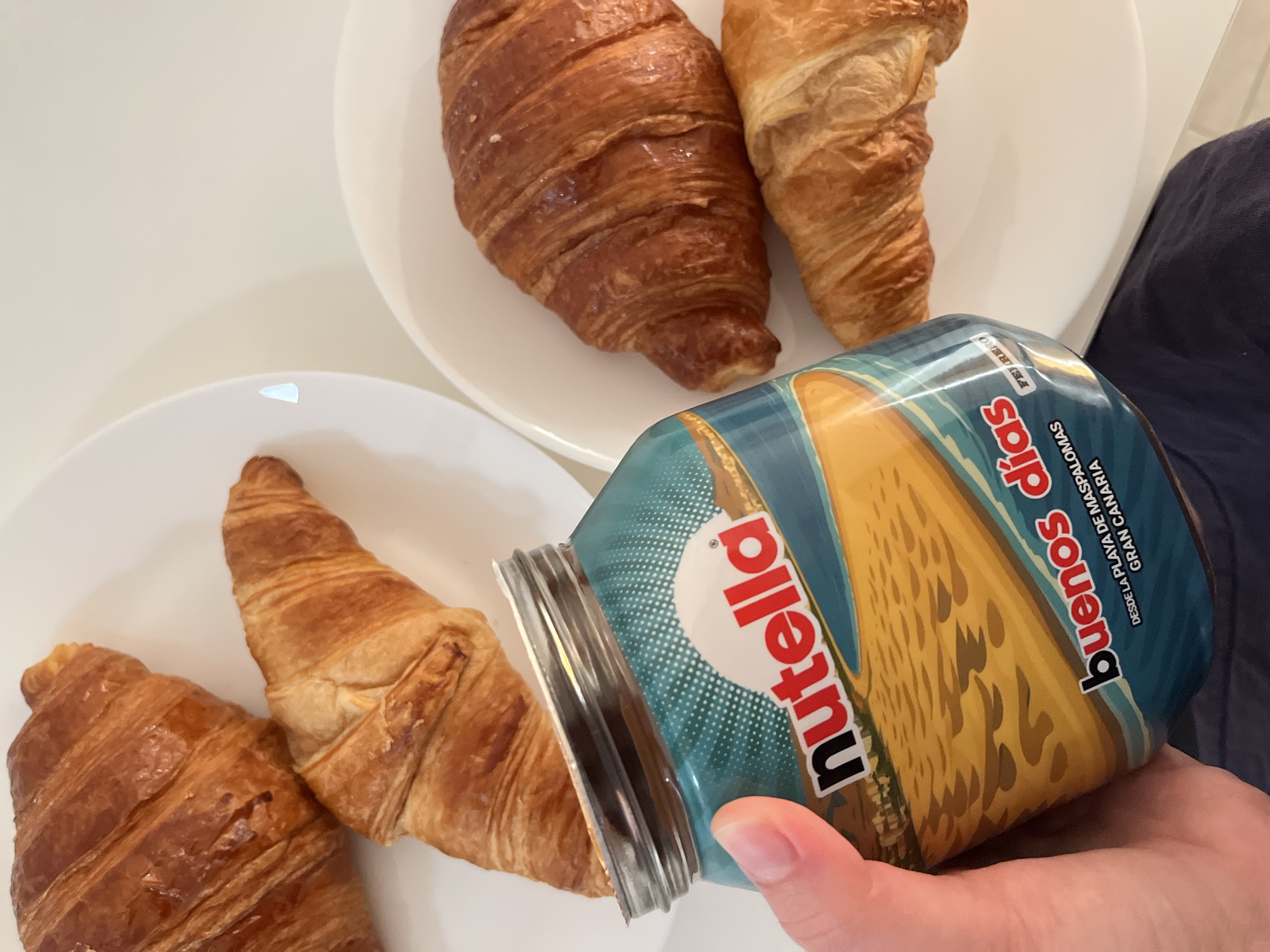 A medium shot depicts a hand holding a jar of Nutella over a plate of croissants. The Nutella jar has a promotional label featuring "Buenos días" and "DESDE LA PLAYA DE MASPALOMAS GRAN CANARIA," indicating the location is related to Mogán, Spain. Two plates are visible, each containing croissants. The croissants vary in color, with some appearing darker and more glazed than others. The background is a plain white surface. The hand is pale and has visible fingernails. The jar is tilted as if pouring Nutella onto the croissants. A portion of a blue shirt is visible in the upper right corner.