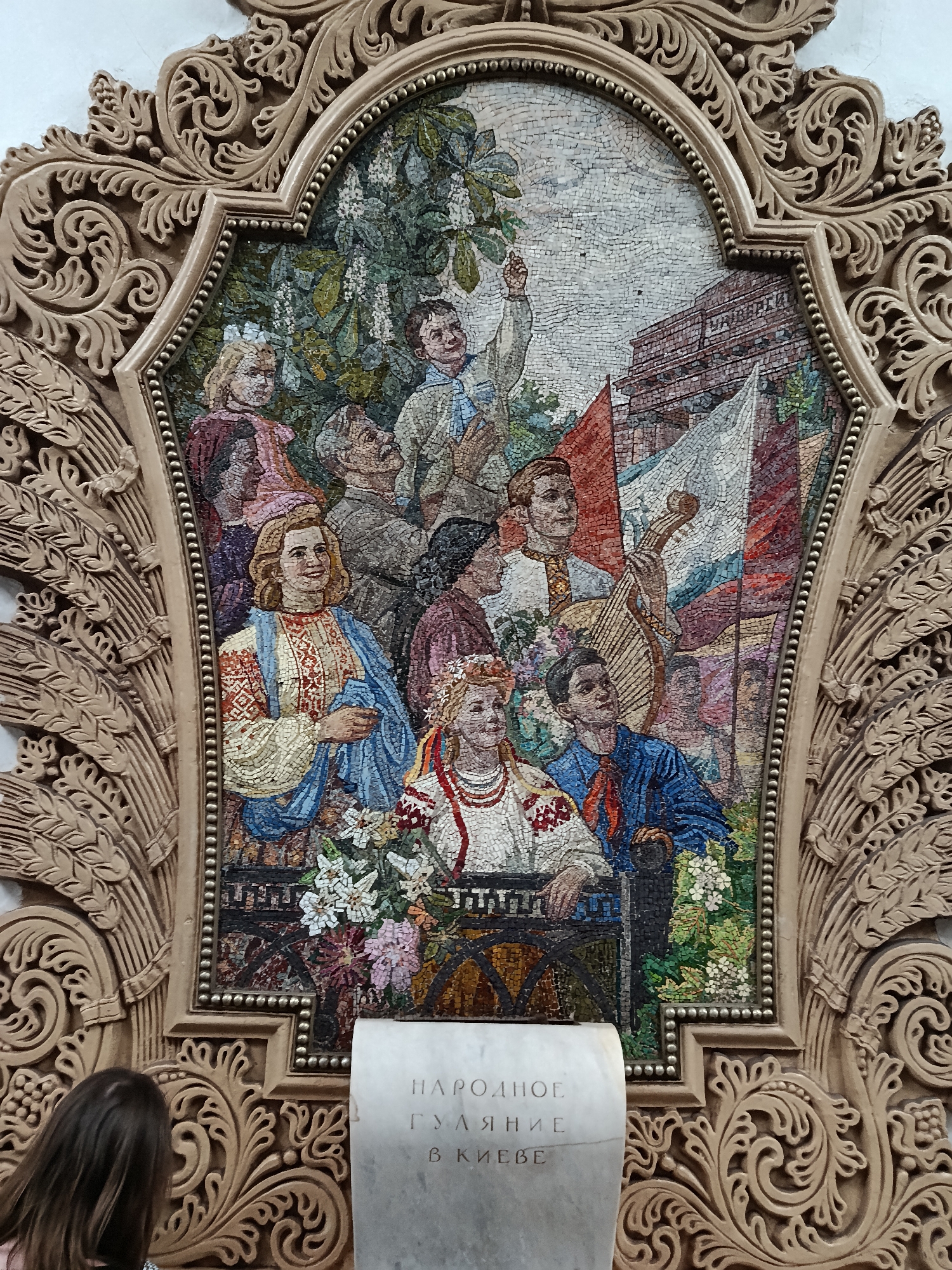 The image shows a mosaic panel within an ornate carved frame. The mosaic depicts a group of people, some in traditional clothing, with flags and a building in the background. The people appear to be celebrating or gathered for an event. In the foreground, to the left, a person's head and shoulders are visible, looking towards the mosaic. Below the mosaic, a stone plaque with text is partially visible. This mosaic is located in the Mayakovskaya Metro Station in Moscow, Russia. The Mayakovskaya station is known for its elaborate mosaics and Art Deco design.