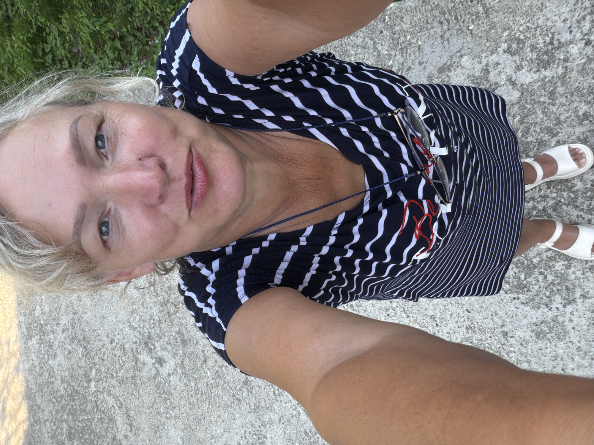 A woman is taking a selfie outdoors. She has blonde hair, blue eyes, and is wearing a striped dress and white sandals. She is standing on a concrete surface, with greenery visible in the background. The image appears to have been taken in Shepsi, Russia.
