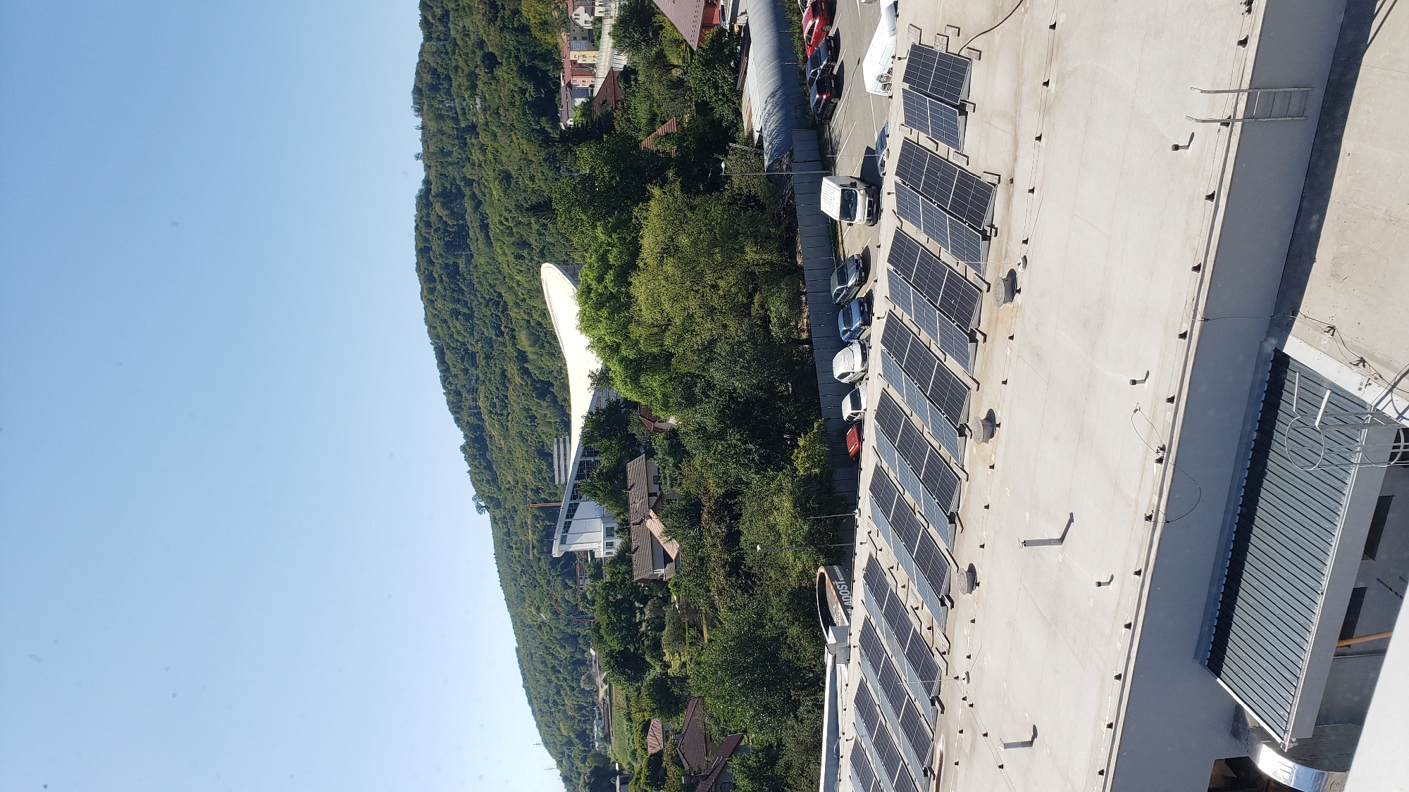 The image depicts a panoramic view of a commercial or industrial rooftop in Prešov, Slovakia, featuring a large array of solar panels. The rooftop itself is a light-colored, flat surface with several circular vents and metallic structures. The solar panels are neatly arranged in rows, angled towards the sky. In the midground, a parking lot is visible with several vehicles, including cars and a white van. Beyond the parking lot, a densely wooded hillside rises, dotted with houses and other buildings. Prominently featured on the hillside is a large, modern white structure with a curved roof, likely a stadium or arena, under a clear blue sky. The overall scene suggests a sunny day in an urban or suburban environment.