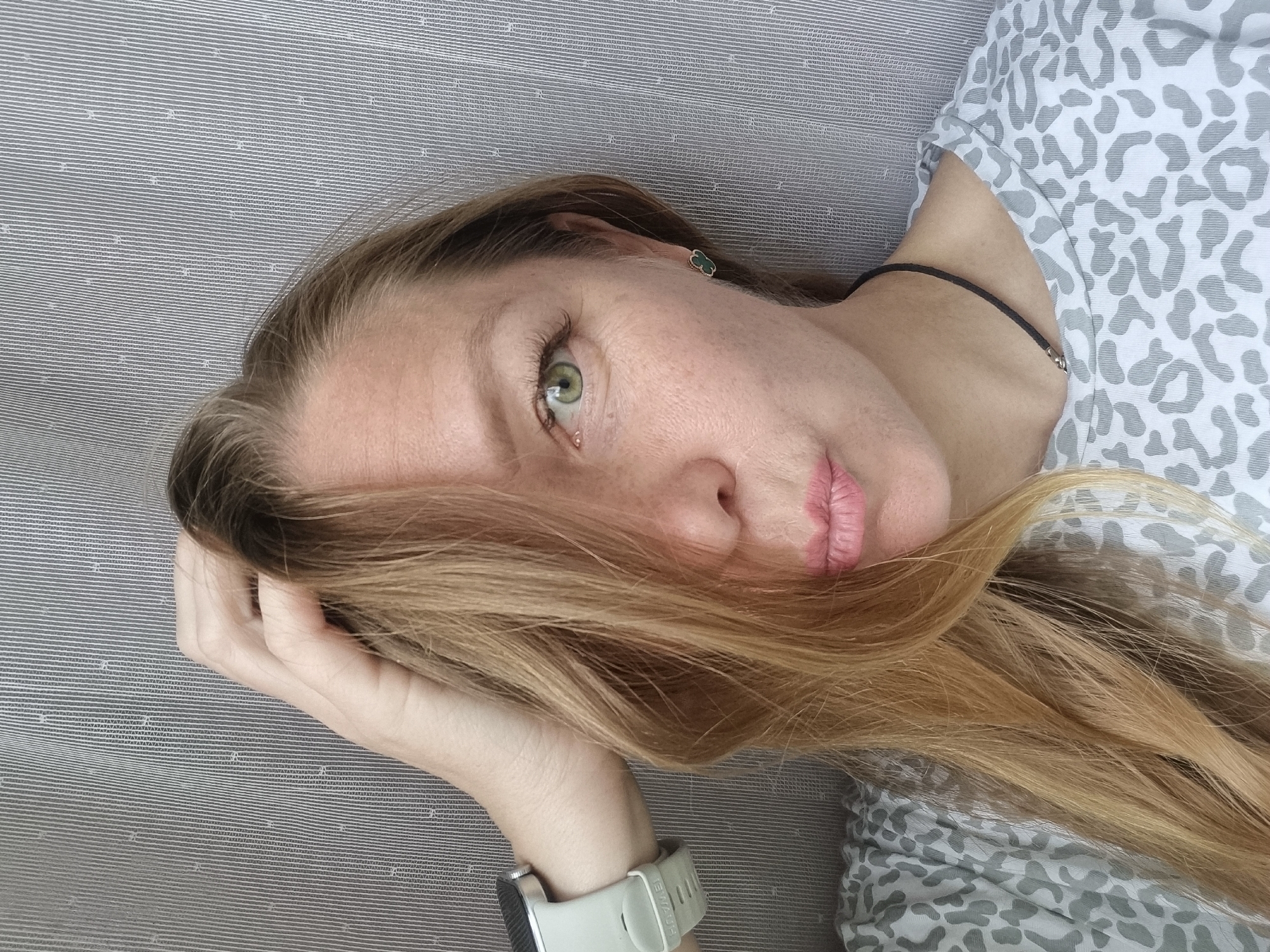 A young Caucasian woman with blonde hair, green eyes, and fair skin with freckles is seen in a close-up, waist-up shot. She is looking directly at the camera with her lips pursed. Her right hand is raised, with fingers touching her hair which is covering part of her face and falling over her left shoulder. She is wearing a light grey t-shirt with a light grey animal print pattern. On her left wrist, she has a light grey smartwatch. She is also wearing a small green earring on her right ear and a thin black necklace. The background is a grey, vertically striped fabric. No specific location in Chaikovsky, Russia, can be identified from the image.