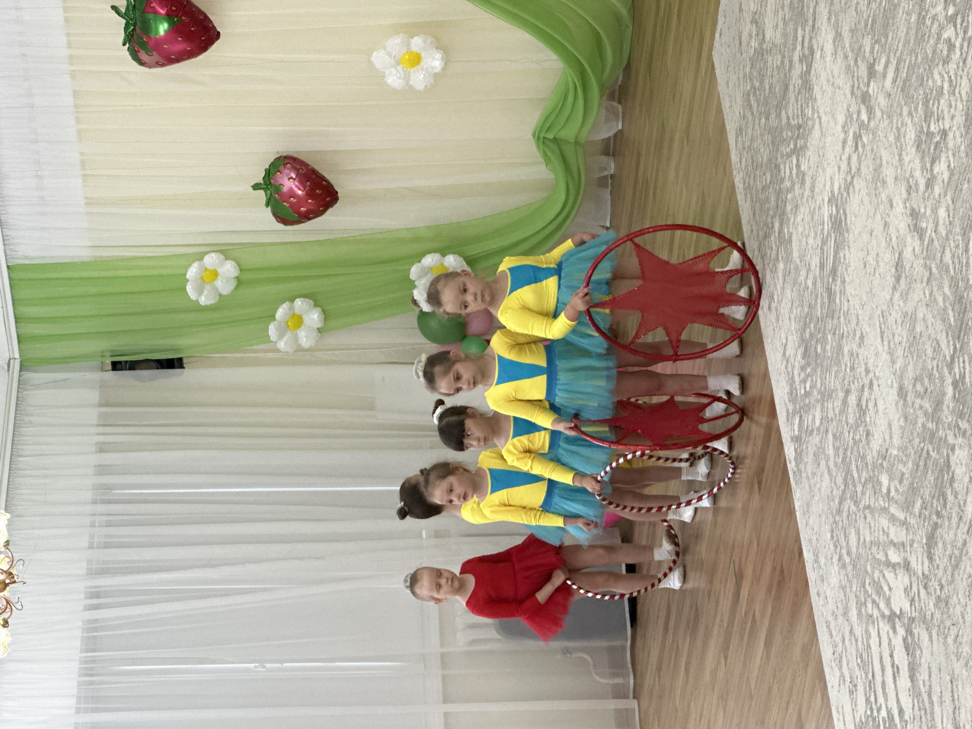 A horizontal indoor shot depicts five young girls standing in a line on a wooden floor. The location is potentially in Chaikovsky, Russia. The first four girls are dressed in identical yellow and blue leotards with matching tutus. The fifth girl, positioned at the front of the line, wears a red dress with a matching tutu. Each girl holds a hula hoop or a star-shaped prop.
Behind the girls is a backdrop of light-colored curtains, adorned with green fabric and artificial daisy flowers. There are also two strawberry-shaped balloons. To the right, a textured white wall is visible. The scene suggests a performance or event, possibly a dance recital or a children's show.
