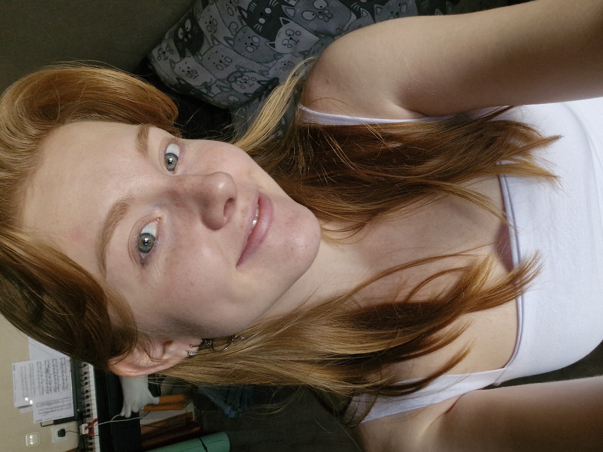 A young woman with reddish-blonde hair and a light complexion is taking a selfie indoors. She is wearing a white tank top and has a small hoop earring in her left ear. Her eyes are a light green, and she has a slight smile on her face. Behind her, a keyboard with sheet music on it is visible, as well as a patterned pillow with cat illustrations. The lighting suggests the photo was taken with artificial light, possibly a flash or overhead light, casting shadows on her face. There is no indication of her location within the city of Yekaterinburg, Russia, or any specific environmental details that would place her there.