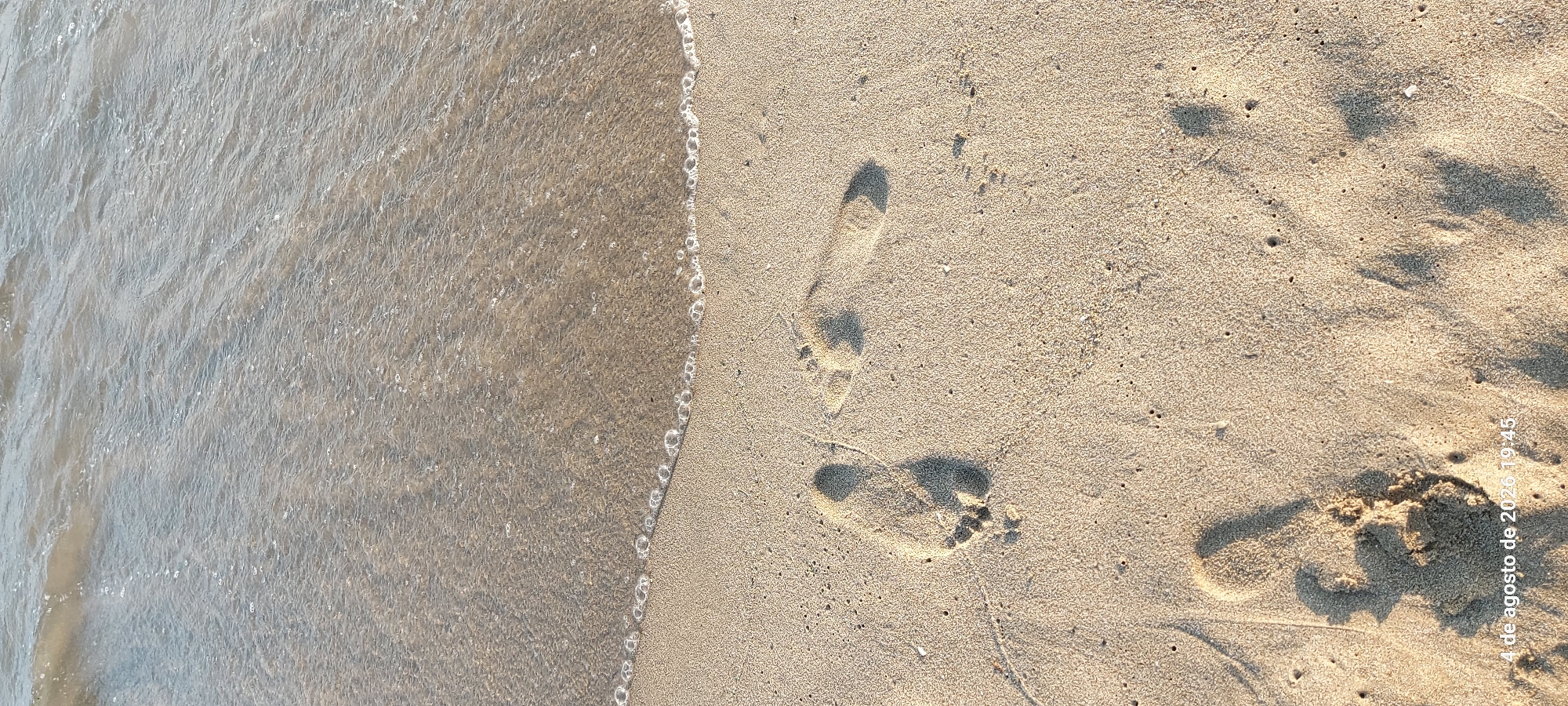 The image captures a close-up view of a sandy beach with the edge of the ocean in the upper portion. Footprints are clearly visible in the sand, indicating human presence. The sunlight casts shadows from the footprints, highlighting their depth and texture. A date and time stamp, "4 de agosto de 2026 19:45", is present in the lower right corner, suggesting the image was taken on August 4, 2026, at 7:45 PM. The environment appears to be a tranquil beach scene at sunset. Without further context or identifying landmarks, pinpointing the exact location within Castelldefels, Spain, is not possible based solely on this image.