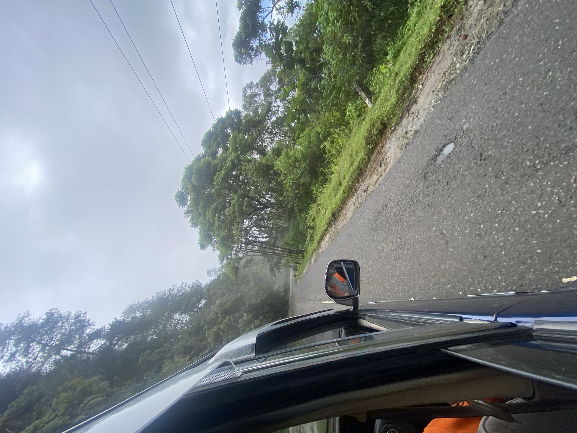 The image shows a view from inside a vehicle driving on a paved road that appears to be in a rural or mountainous area, likely in Palopo, Indonesia. The road is lined with lush green vegetation and trees on the right side, with a steep, forested incline on the left. The sky is overcast and hazy, suggesting a cloudy or misty condition. A utility pole with power lines is visible in the upper right portion of the image. The side mirror of the vehicle reflects a person wearing an orange garment, possibly the driver or a passenger. The road surface shows signs of wear, with some potholes and loose gravel. The overall scene depicts a journey through a natural landscape.