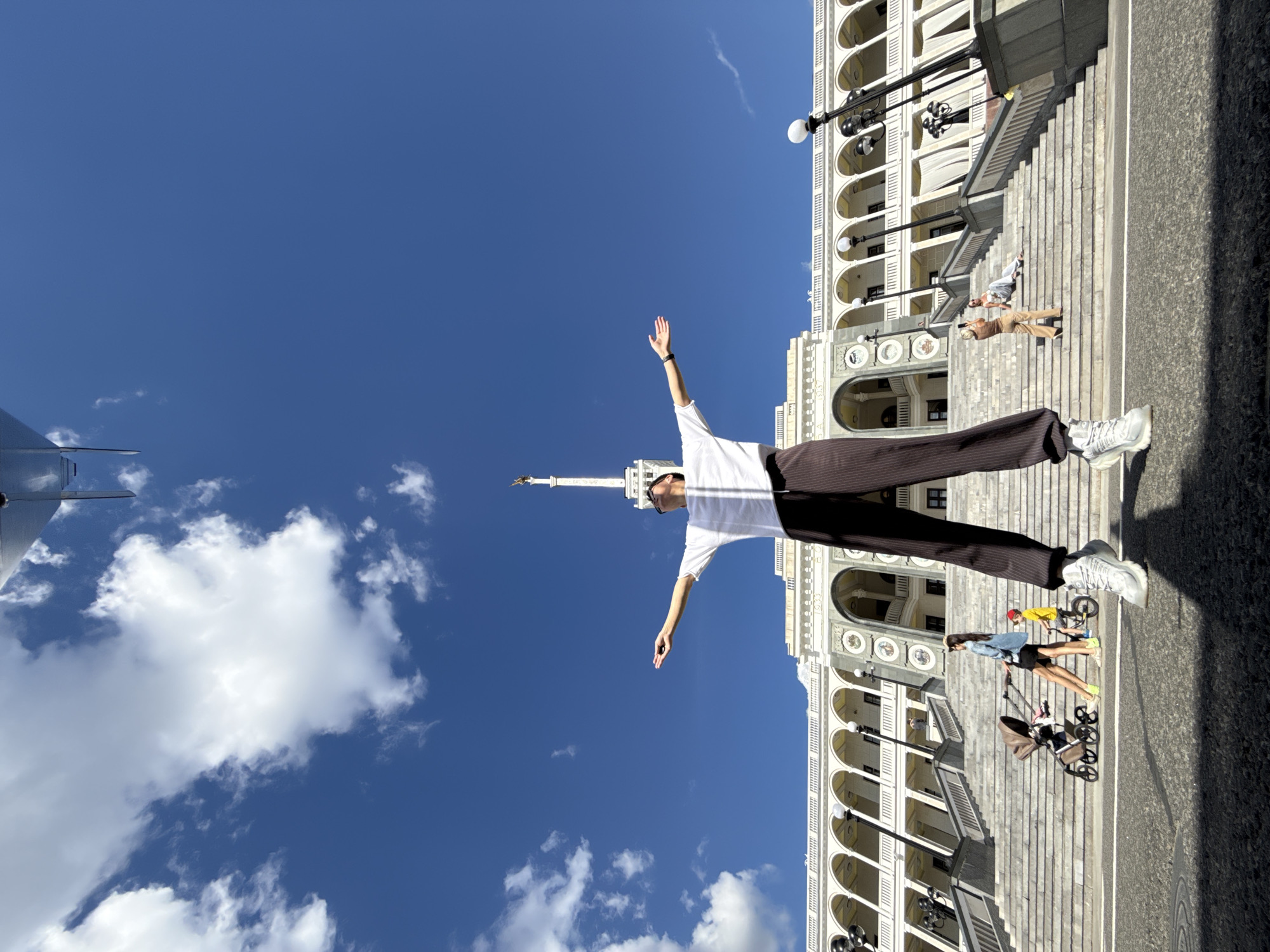 A low-angle, wide-shot photograph taken outdoors on a sunny day showcases the Khimki River Station in Khimki, Russia. The station, a grand structure with a colonnaded facade and a prominent central spire topped with a golden eagle statue, serves as the backdrop. In the foreground, a person stands with their arms outstretched, looking upwards and wearing a white t-shirt, dark striped pants, and white sneakers. They are positioned on a wide, paved area in front of a large set of stairs leading up to the station. Several other individuals are visible on the stairs, including a woman with a stroller and a child on a bicycle, and another woman taking a photo of a person sitting on the stairs. The sky is a vibrant blue with scattered white clouds, and a portion of an unidentified metallic object, possibly part of an aircraft, is visible at the top of the frame.
