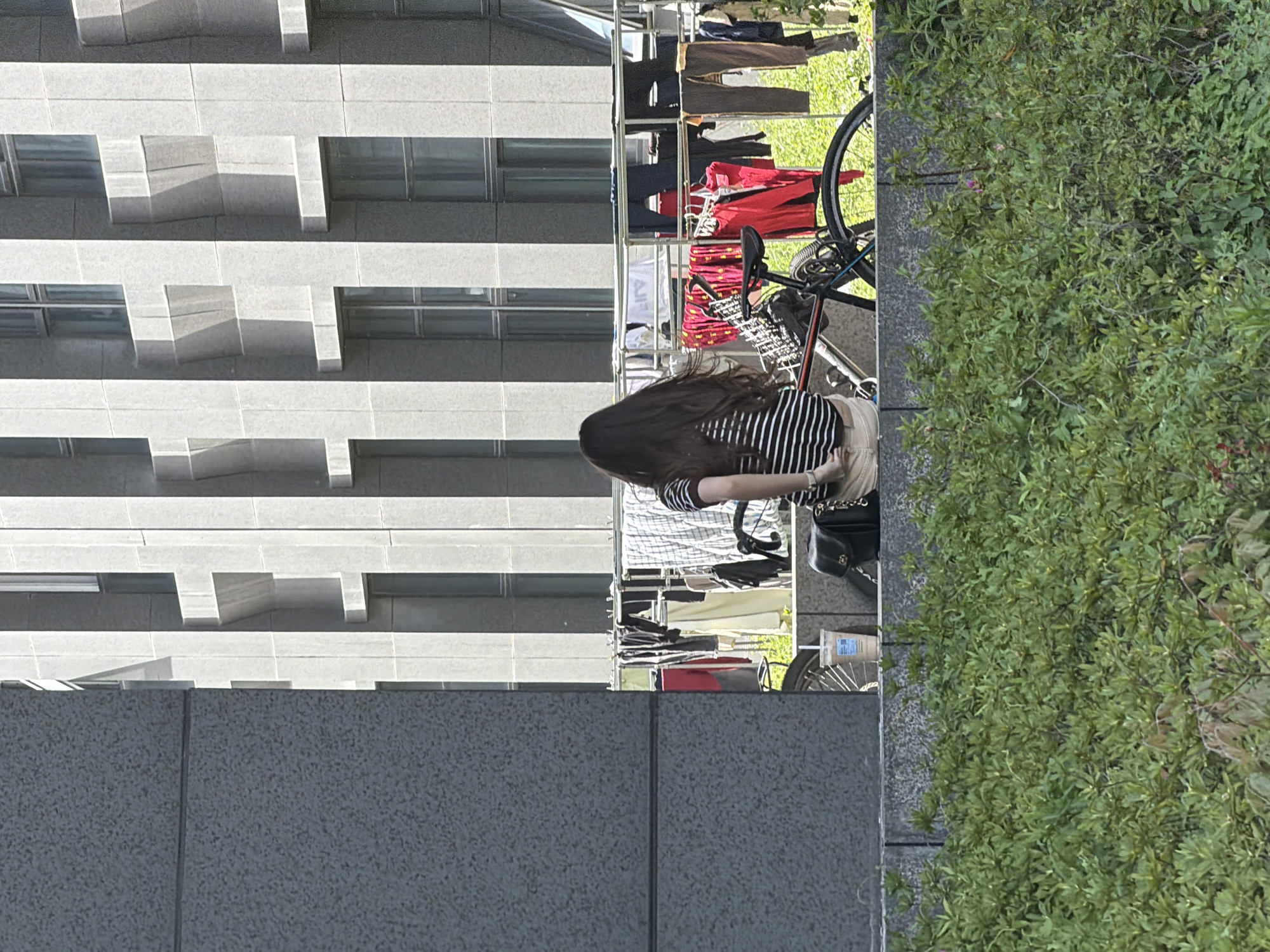 A medium, eye-level shot captures a young woman with long brown hair wearing a black and white striped shirt and khaki pants. She stands next to a bicycle parked on a sidewalk, with a clothing rack filled with hanging clothes behind her. The background features a modern, grey building with horizontally layered rectangular window designs. Dense green vegetation lines the edge of the sidewalk, bordering the building. The scene is located in Jiangning District, China.