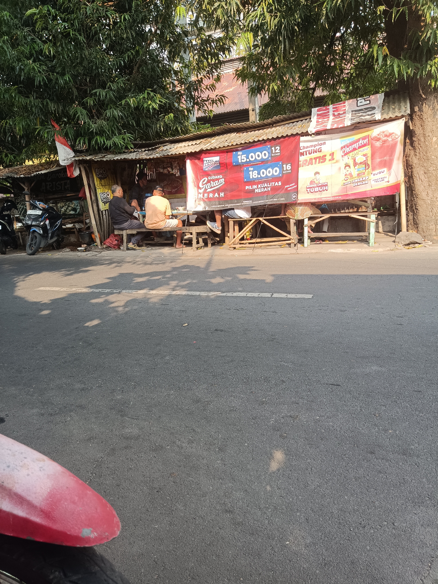 The image depicts a street scene in Jember, Indonesia, with a small roadside stall and several individuals. In the foreground, the asphalt road is visible with shadows cast by trees. A portion of a red motorcycle fender is also present in the bottom left corner.
Behind the road, a simple, open-air stall is set up. It has a corrugated metal roof and is supported by wooden structures. Several banners are hung along the front of the stall. One banner displays prices in Indonesian Rupiah, showing "15.000" and "18.000". Another banner features the brand "Champion" and text in Indonesian, possibly related to health or supplements, mentioning "GRATIS 1" and "ENERGY UNTUK TUBUH".
Three men are seated at a wooden table in front of the stall. One man, wearing a black shirt, is facing away from the camera, while another man in an orange shirt is seated next to him, also facing away. A third man, wearing a black shirt and a cap, is visible in the background within the stall area.
To the left of the stall, a blue motorcycle is parked. The environment is characterized by the presence of lush green trees providing shade, and the general atmosphere suggests a typical street-side establishment in Indonesia. The time of day appears to be late afternoon, judging by the long shadows.