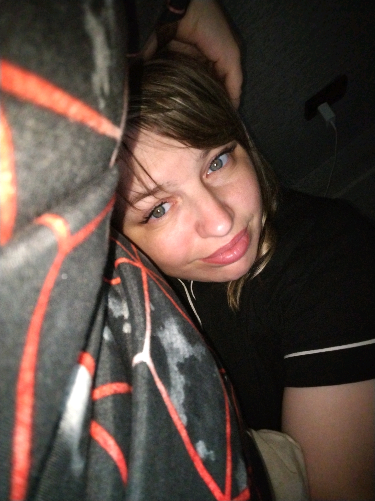 The image is a selfie of a woman with light brown hair and blue-green eyes. She is wearing a black t-shirt with white piping. Her left arm is resting on a dark grey blanket with a red geometric pattern. Her right hand is on her head. The background is dark and appears to be indoors, possibly a bedroom. A small black object, possibly a phone, is visible in the upper right corner of the image, connected to a white wire. The woman has pink lipstick on and appears to be looking at the camera. There is no visible information in the image to indicate the location in Yaroslavl, Russia.