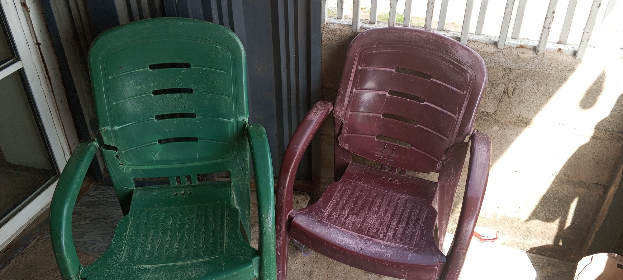 This media file captures an outdoor scene in Jalingo, Nigeria, featuring two plastic chairs. The chair on the left is a vibrant green, while the one on the right is a deep burgundy. Both chairs appear to be slightly weathered, showing signs of dust accumulation on their surfaces, suggesting they have been exposed to the elements. The green chair has visible damage on its armrest, indicating wear and tear.
The chairs are positioned next to a wall constructed of concrete blocks, which has a rough, textured finish. Behind the chairs, a metal corrugated structure is visible, likely part of a building or enclosure. In the background, a white metal railing or fence can be seen, separating the area from what appears to be an outdoor space with some vegetation or ground cover. Sunlight is casting distinct shadows on the wall and ground, indicating it is daytime. The shadows are long and sharp, suggesting the sun is either low in the sky or angled at a specific point, possibly late afternoon or early morning.
The scene is set in an outdoor environment, potentially a patio, veranda, or yard. There are no people visible in the image, and no specific activity is taking place. The overall impression is one of stillness and quiet, with the focus solely on the two chairs and their surroundings. The light and shadow play adds a dynamic element to the static subjects. No text is visible in the image.