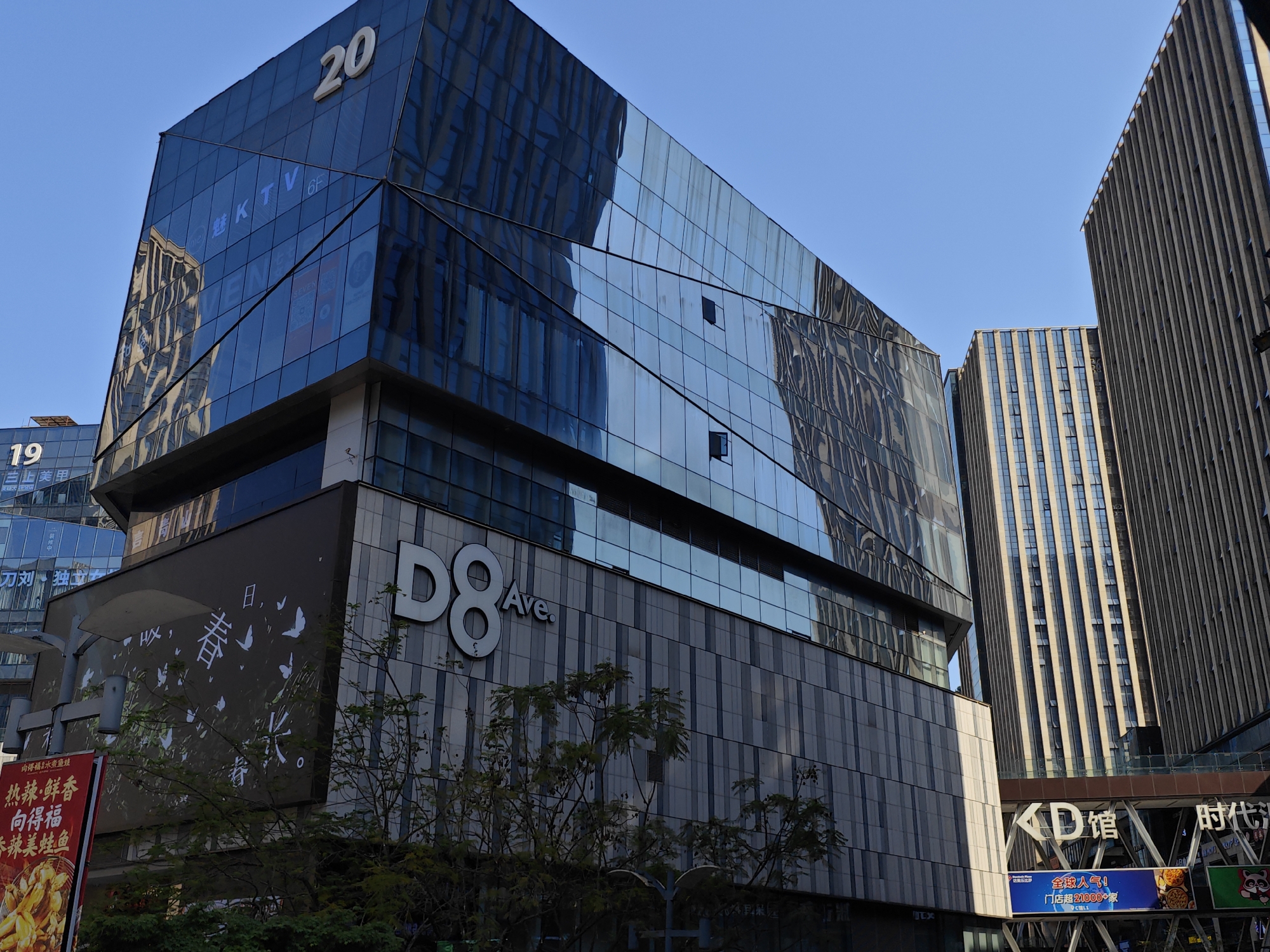 This image depicts a bustling urban scene in Yuzhong District, China, characterized by modern architecture under a clear blue sky. The central focus is a large, multi-story commercial building with an angular, reflective glass facade that mirrors the surroundings. A prominent white number "20" is visible near the top, while "KTV" and "6F" are displayed in blue lighting on the glass. The lower section of this building features grey textured panels with large white lettering that reads "D8 Ave." A dark panel below the glass also displays white Chinese characters, including "春" (spring) and "日" (day), accompanied by butterfly designs.
Adjacent to the main building on the left, another structure with a glass facade is partially visible, bearing the number "19". In the background and to the right, several other tall, contemporary skyscrapers with vertical patterned facades rise, indicating a dense urban environment.
At ground level, partially obscured by trees, various commercial advertisements are present. A red sign prominently advertises "向得福水煮鱼蛙" (Xiang Defu Boiled Fish and Frogs) with phrases like "热辣鲜香" (Hot, Spicy, Fresh, Fragrant). Further to the right, beneath an elevated structure, signs for "KD馆" (KD Hall) and "时代" (Times) are visible, along with a blue banner proclaiming "全球人气!" (Global Popularity!) and "门店超21000+家" (Over 21000+ stores). The bright, clear sky and distinct shadows suggest a sunny daytime setting. No individuals are directly visible, but the extensive commercial signage implies a lively retail and entertainment district.