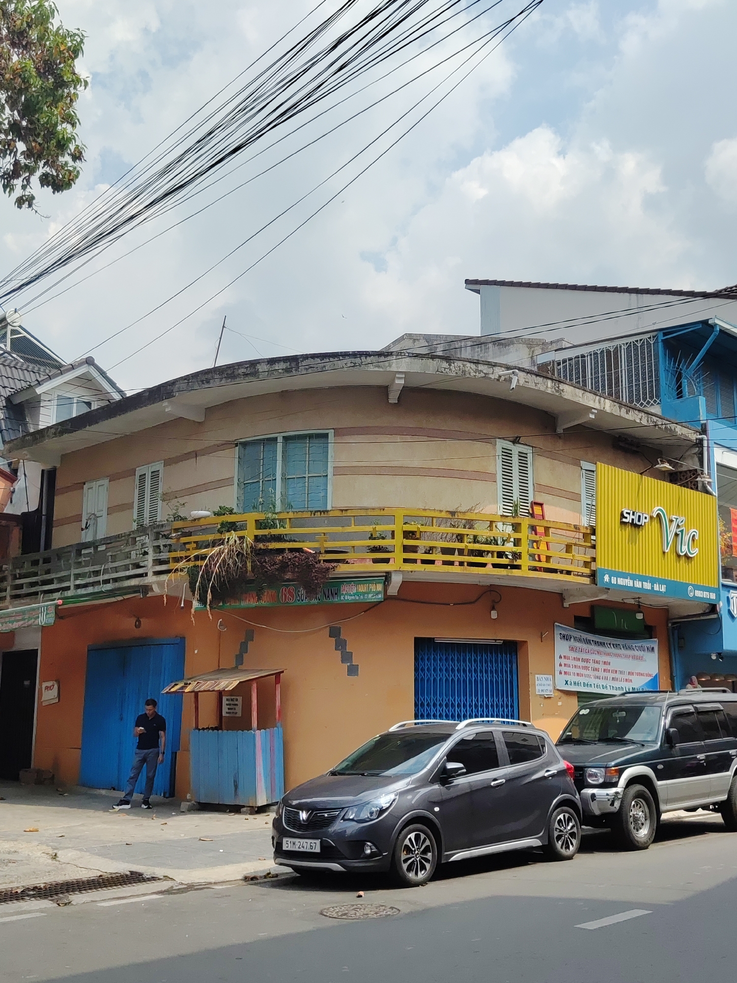 The image shows a street scene in Da Lat, Vietnam. A two-story building with a rounded upper section and a yellow balcony is the focal point. The building houses a shop named "Shop Vic" located at 68 Nguyen Van Troi, Da Lat. An individual is standing near a blue-painted stall, looking at a phone. Two cars are parked along the side of the road: a gray hatchback and a dark SUV. Electrical wires are visible overhead, intersecting with the sky and partially obscuring the building. Signage with Vietnamese text is affixed to the building's facade.