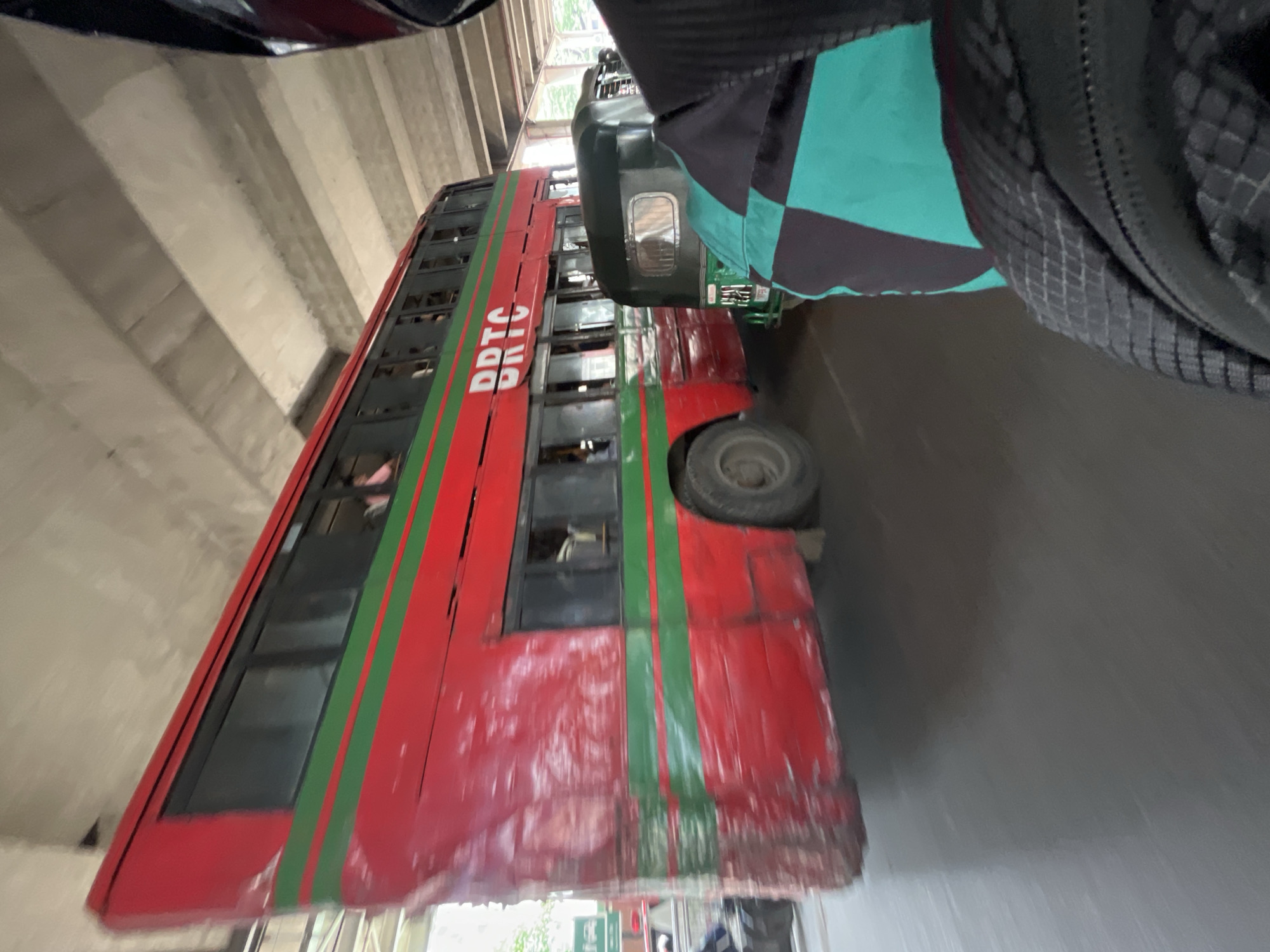 The image shows a red double-decker bus, labeled "BRTC" in white letters, under an overpass. The bus has a green stripe running vertically along its side. The bus is near the side of the road, as a gray pavement is visible. A part of a person wearing a green and black jacket is visible in the upper right of the picture. Part of another vehicle can be seen at the top left of the image. The overpass is made of concrete. Based on the BRTC buses, the location is likely Dhaka, Bangladesh.