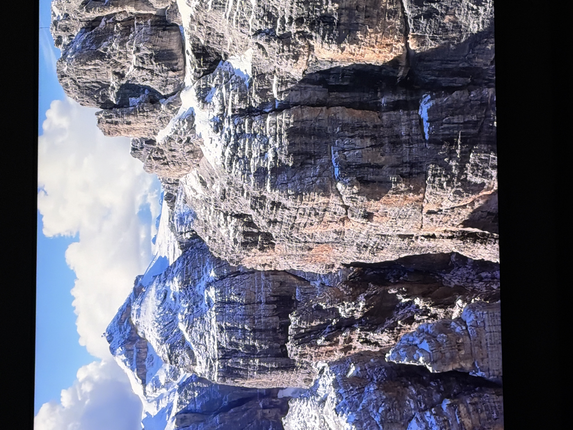 The image displays a majestic mountain range under a bright blue sky with scattered white clouds. The mountains are rugged and rocky, with patches of snow clinging to their upper slopes and crevices. The sharp contrast between the dark rock and the white snow creates a dramatic visual effect. The scale of the mountains is imposing, with sheer cliffs and jagged peaks. The scene evokes a sense of wilderness and natural beauty. There are no people or man-made structures visible in the image. This image is not located in the city of Chirchik, Uzbekistan, as Chirchik is a city and not known for its mountain ranges.