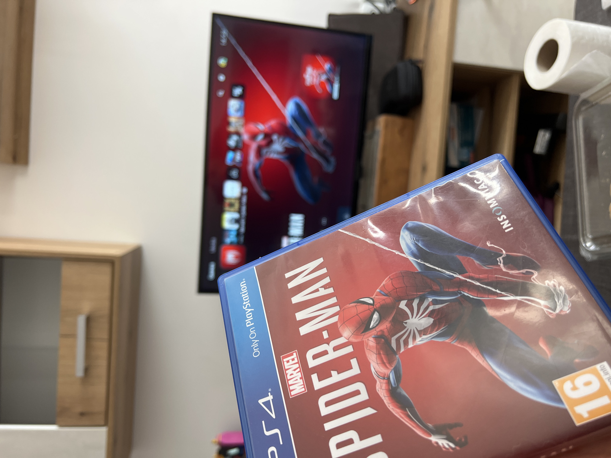 The image shows a PlayStation 4 game case for "Spider-Man" in the foreground. Behind the game case, there is a television displaying what appears to be the same "Spider-Man" game. The environment includes a light-colored cabinet to the left of the TV. To the right of the TV, there is a shelving unit, and objects such as a roll of paper towels and a container are visible. The location is not discernible as Burgas, Bulgaria based solely on the visual data provided.