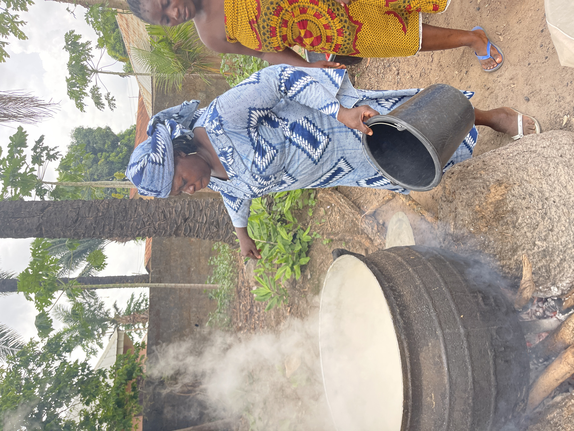 The image shows a woman in a blue patterned dress and headwrap pouring from a black bucket into a large, dark pot over an open fire. Smoke is rising from the pot. Another person, wearing a yellow and red patterned dress, is partially visible standing behind the woman. The scene appears to be outdoors, possibly in a rural area of Akwanga, Nigeria, given the presence of vegetation, bare ground, and basic structures in the background.
