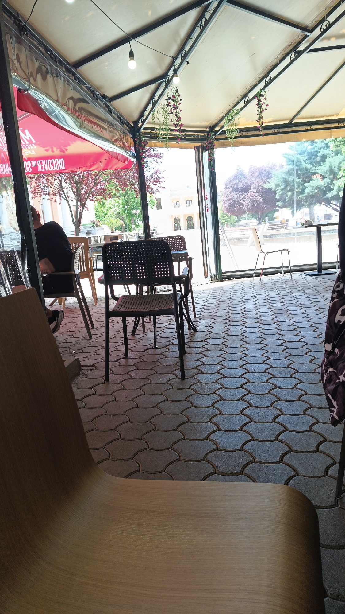 The image shows the interior of an outdoor café in Karnobat, Bulgaria. The seating area is covered by a semi-permanent structure with a light-colored fabric roof supported by a black metal frame decorated with artificial hanging plants. The floor is paved with grey, interlocking pavers. A variety of chairs are visible: dark plastic chairs with perforated backs, light wood chairs, and a modern wooden chair in the foreground. At least one person is seated at a table, visible partially obscured by a chair in the background. Through the open sides of the café structure, there are glimpses of trees, a building, and outdoor furniture, suggesting a public space or park setting. A red awning is partially visible on the left.
