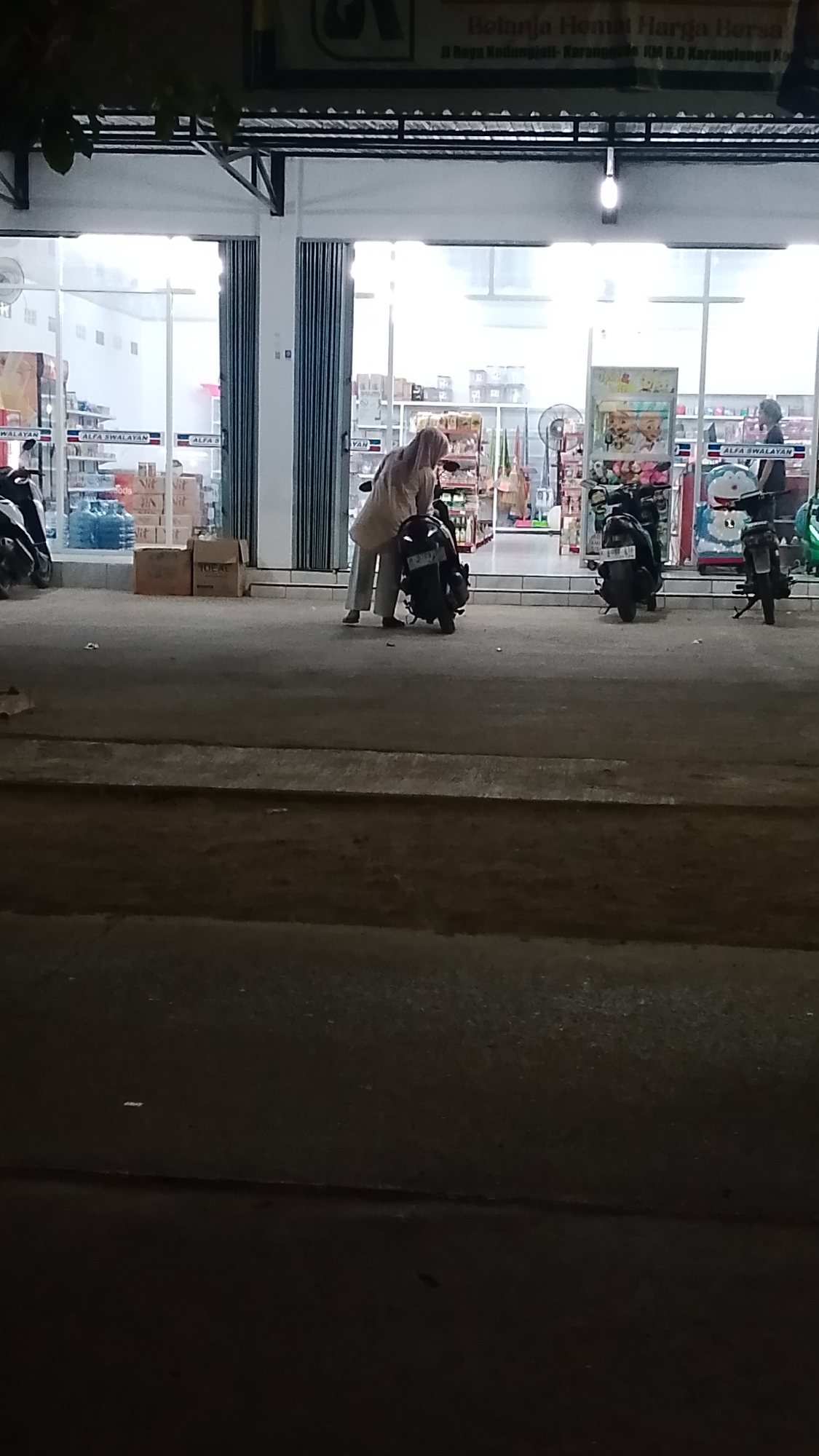 The image depicts a street scene in front of a minimarket in Karanglangu, Indonesia, likely at dusk or night due to the lighting conditions. In the foreground, a dark asphalt road is visible, with a raised concrete curb separating it from the sidewalk.
The minimarket, identified by signage that appears to include "ALFA SWALAYAN," has a wide glass storefront with folding metal doors partially open. Inside, shelves stocked with various products are illuminated. A person wearing a light-colored headscarf and loose-fitting clothing is crouched beside a dark-colored scooter parked directly in front of the minimarket entrance. Their posture suggests they might be adjusting something on the scooter or interacting with its storage.
Two other scooters are also visible parked in front of the minimarket. One is to the left of the central figure, and another is further to the right, near a display featuring cartoon characters. The overall environment is that of a commercial area with pedestrian and vehicular access, common in Indonesian cities. The presence of multiple parked scooters indicates a common mode of transportation in the area.