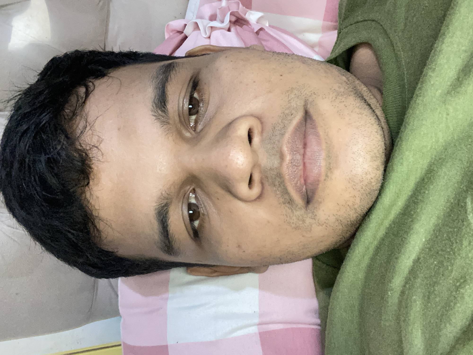 A close-up, overhead, selfie-style shot shows the face of a South Asian man with dark hair, brown eyes, and light stubble. He is lying down with his head on a pillow, which has a pink and white checkered pattern. A portion of a light-colored couch or chair is visible behind his head. He is wearing a green t-shirt, which is folded and wrinkled. The lighting is natural and diffused, with no strong shadows. The background is out of focus. The setting appears to be indoors, possibly a bedroom or living room, in a location identified as Unknown, Unknown.