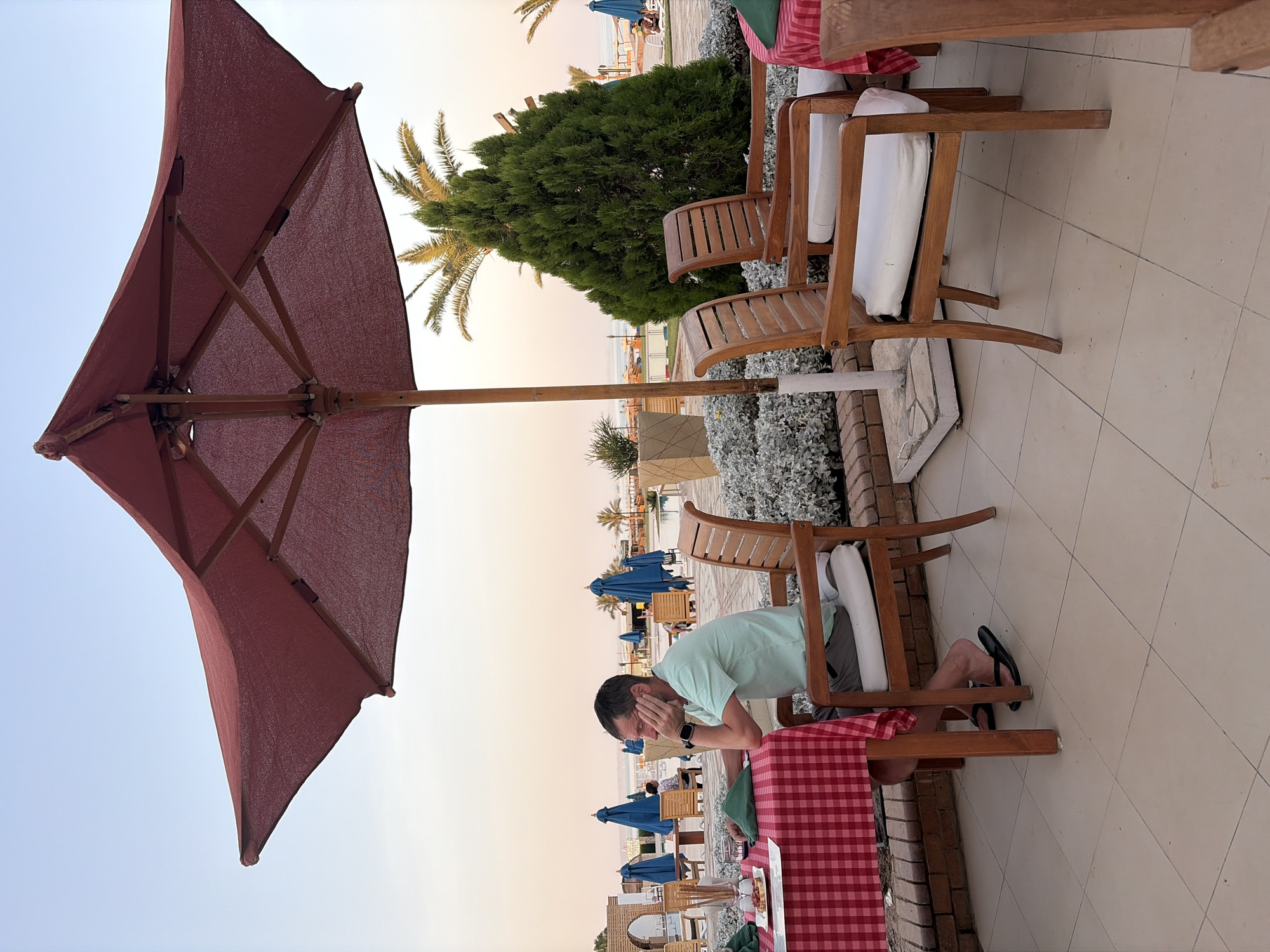 A man is sitting at a table covered with a red and white checkered tablecloth in an outdoor restaurant at the Mercure Hurghada, Egypt. The man, wearing a light green t-shirt and flip-flops, rests his head in his hands, appearing to be deep in thought. A plate with food and a glass of water are on the table in front of him. Wooden chairs with white cushions are arranged around the table. In the background, a large red umbrella provides shade, and further beyond, a swimming pool and the Red Sea are visible. Palm trees and lush green bushes dot the landscape, suggesting a tropical resort setting. The sky is a pale orange, indicating either sunrise or sunset.