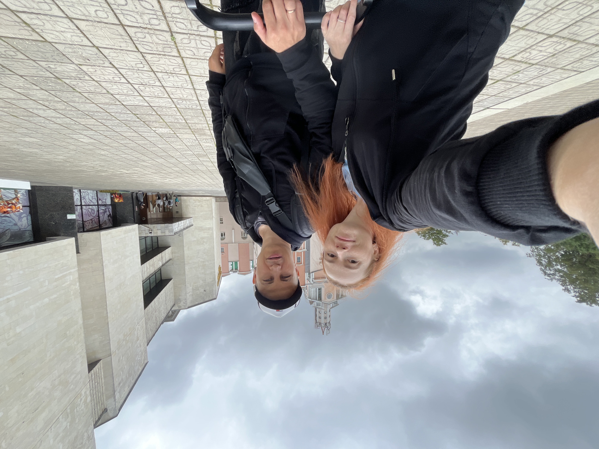 A couple is taking a selfie in front of the State Tretyakov Gallery, located in Moscow, Russia. The woman, on the left, has long, reddish-orange hair and is wearing a black zippered hoodie. The man, on the right, is wearing a black baseball cap, a black hoodie, and a black crossbody bag. They are standing on a paved plaza with a tiled pattern. Behind them, the building has a facade made of light-colored stone blocks and large windows. The sky is overcast with gray clouds.