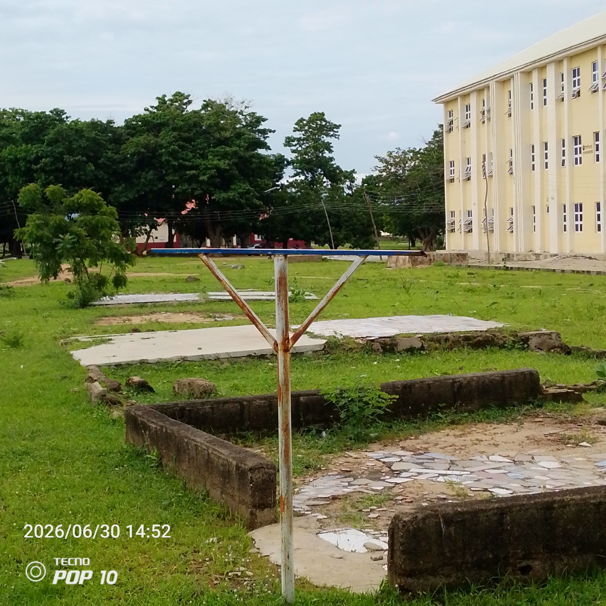 The image depicts an outdoor scene in Jalingo, Nigeria, featuring a large, light-yellow building with multiple floors and numerous windows. In the foreground, a rusty metal pole with a V-shaped support structure is visible, positioned on a patch of grass. Surrounding the pole are low, dark-colored concrete walls, forming a rectangular enclosure. Beyond this, there are paved areas with scattered stones and patches of grass. A line of mature green trees forms a backdrop, with a cloudy sky overhead. The timestamp "2026/06/30 14:52" and the "TECNO POP 10" logo are superimposed on the lower left corner of the image.