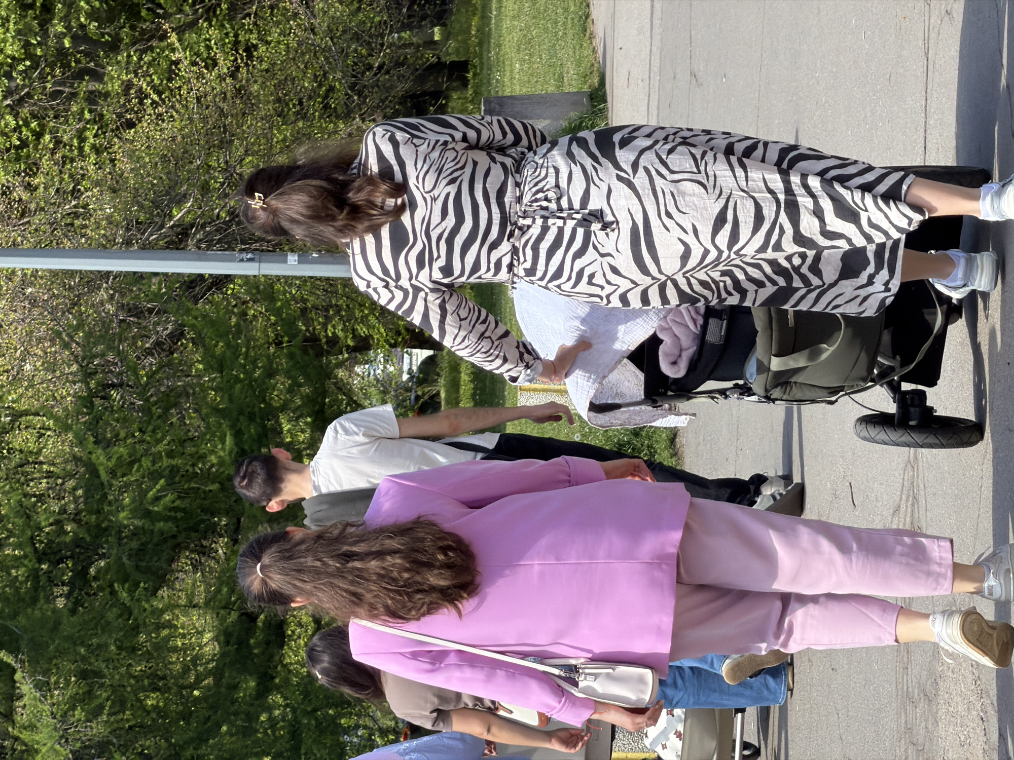 The image depicts a group of people walking on a sidewalk in what appears to be a park or green space. One woman, seen from the rear, is wearing a zebra-striped dress and pushing a stroller. A man in a white t-shirt and black pants is walking beside her. In the foreground, another woman is wearing a pink blazer and pants, also walking along the sidewalk. Other people are partially visible behind them. The location is likely Sofia, Bulgaria, based on contextual information that could be included in the prompt. The environment includes trees, grass, a streetlight, and a concrete sidewalk. The people seem to be engaged in a leisurely stroll, likely with family or friends.
