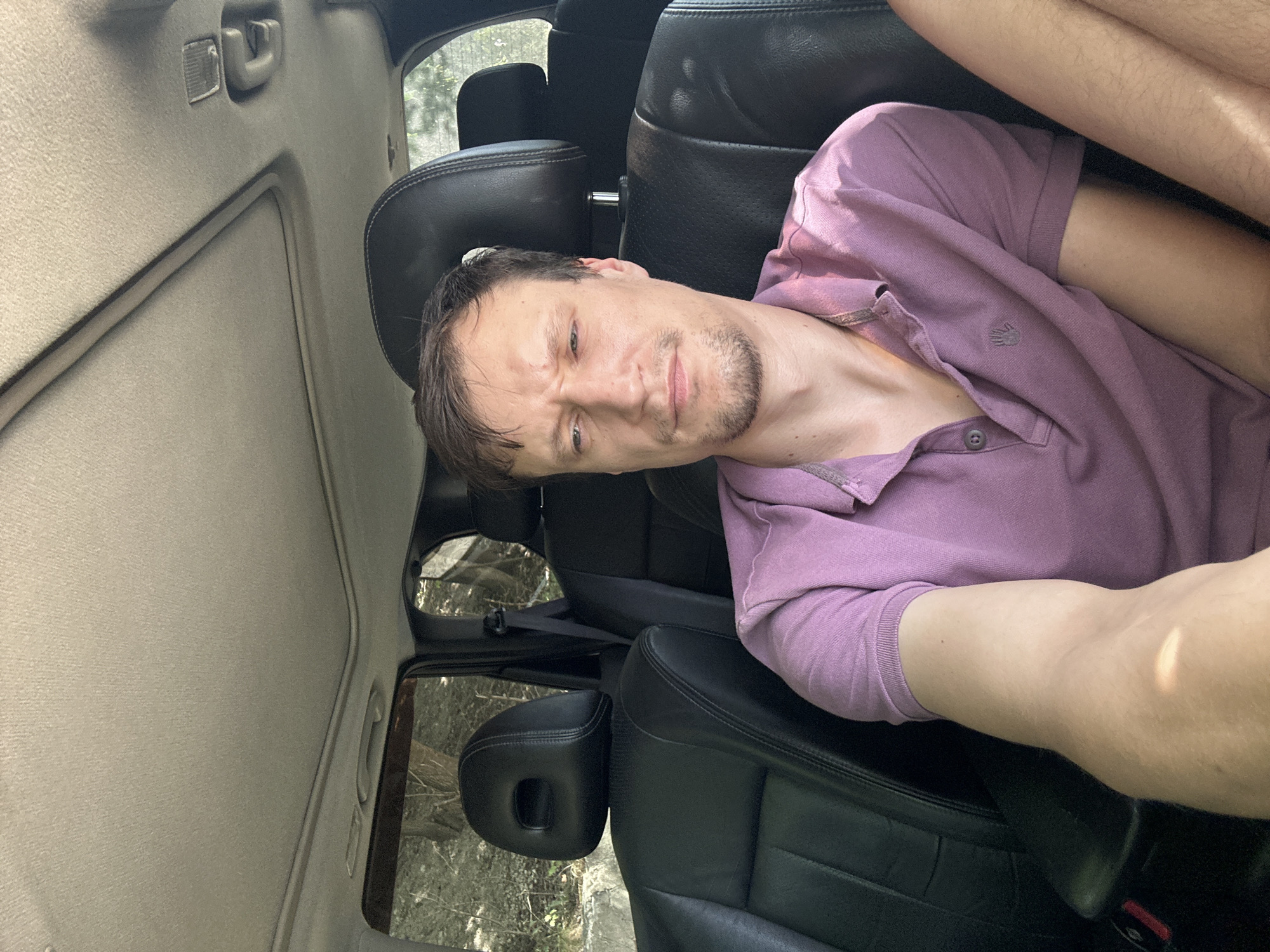A man is sitting in the passenger seat of a car. He is wearing a purple polo shirt. The car's interior is visible, with black leather seats and a sunroof. Outside the car, trees and foliage can be seen. The image appears to be taken during the daytime, with sunlight casting shadows. The location in the city of Makiv, Ukraine is not discernible from the image.