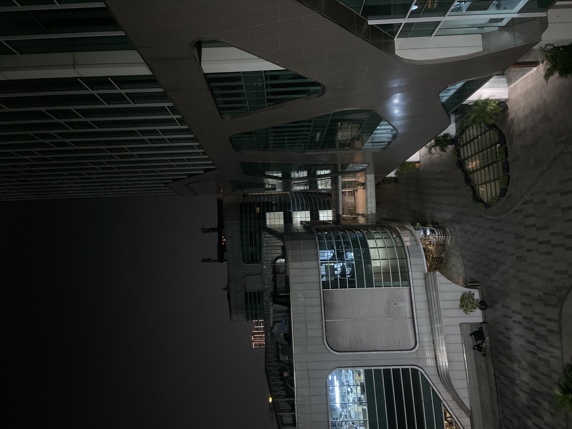 An eye-level, medium shot, taken at night, shows a contemporary building in North Jakarta, Indonesia. The building features a modern design with rounded corners, glass windows, and geometric shapes. A covered, tiled, outdoor walkway connects different parts of the structure. The walkway has several openings that reveal illuminated interiors. In the lower right corner, two people are visible near a potted plant on the walkway. The sky is dark and partially visible on the left side of the image. The surrounding area is dimly lit, and the overall atmosphere is modern and urban.