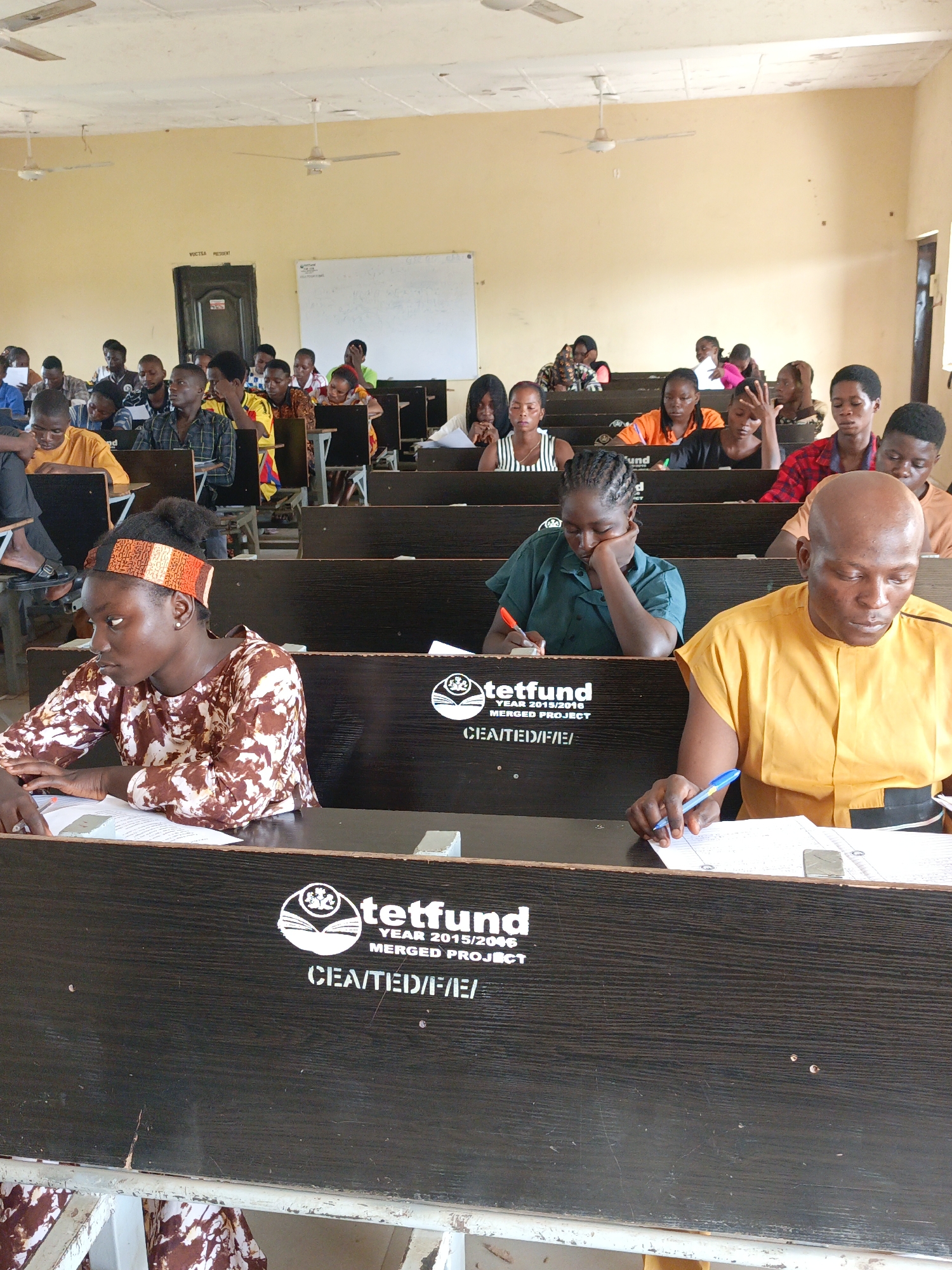 The image shows a classroom in what appears to be a school or university setting. Students are seated at desks with "tetfund" logos on them, suggesting this is a project funded by the Tertiary Education Trust Fund. Most are focused on papers in front of them, potentially taking a test or exam. There are 30+ individuals of apparent African descent in the classroom with a whiteboard and an open door. The classroom walls are light yellow and there are ceiling fans. Based on the text "VOCTRA PRESENT" it is likely the vocational training center in Akwanga, Nigeria.