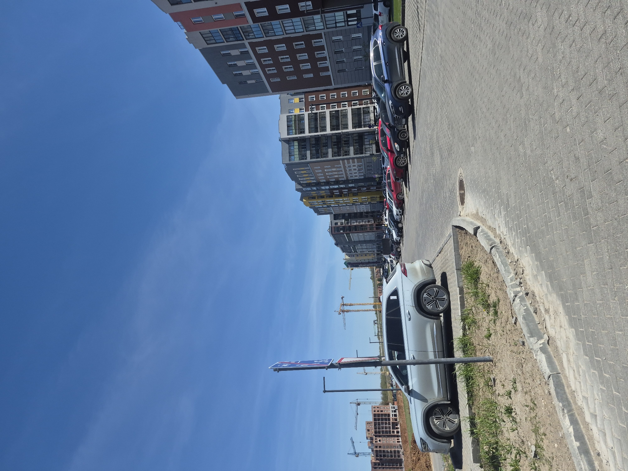 The image shows a street in Pervomaysky, Russia. Several modern apartment buildings line the street, and cars are parked along the curb. In the distance, construction cranes indicate ongoing development. The sky is clear and blue. A light-colored SUV is parked in the foreground, partially blocking a street sign. The street is paved with grey brick and has a raised curb separating it from a narrow strip of unpaved ground with sparse vegetation.