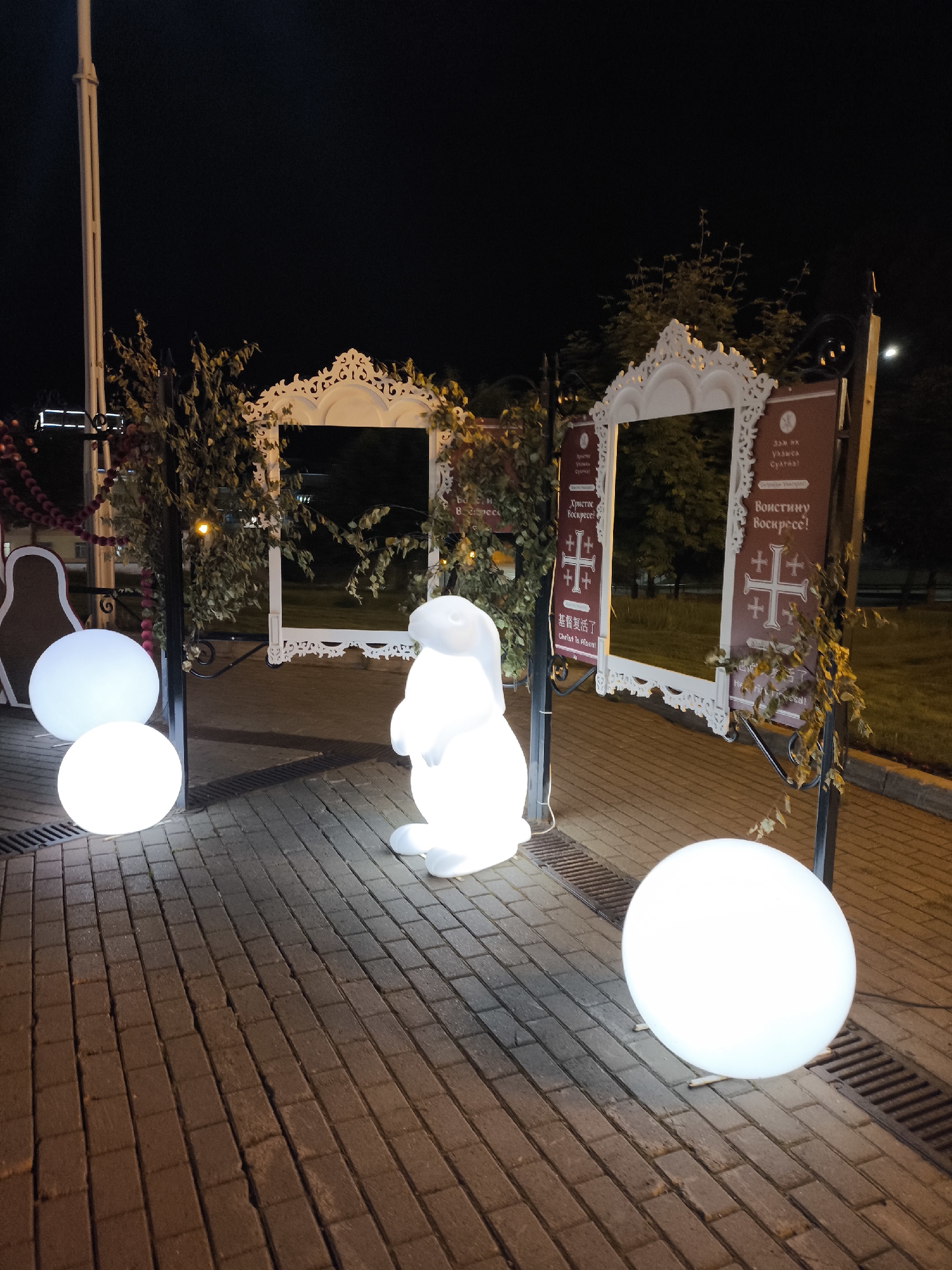 This image depicts an outdoor nighttime festive display on a paved pedestrian area in Izhevsk, Russia. The scene is illuminated by the objects within the display and some distant ambient light, with a dark sky overhead.
In the foreground, a paved brick walkway stretches diagonally from the lower left to the upper right. Several white, illuminated spherical lights are positioned on the ground; two are grouped on the left side, and one larger sphere is on the right. To the immediate right of the two left spheres, a prominent white, glowing, rabbit-like figure stands upright.
Behind these illuminated objects, a decorative installation frames the view. It consists of two large, ornate, white-framed structures, resembling archways or oversized picture frames, positioned side-by-side. The frames feature intricate, carved scrollwork details along their tops and bottoms. Greenery, possibly artificial vines, is draped over the black metal supports holding these frames, with some additional autumn-colored foliage visible.
Attached to the supports on either side of the frames are rectangular signs with a dark red background and white text. The sign on the left frame displays the greeting "Христос Воскресе!" (Christ is Risen!) in Russian, and "基督复活了" (Christ is Risen!) in Chinese, accompanied by a white cross symbol with four smaller crosses in its quadrants. The sign on the right frame features "Воистину Воскресе!" (Indeed He is Risen!) in Russian, also with the same cross symbol, and "Christ is Risen!" in English and "基督复活了" in Chinese below it. Partially visible text above the main greeting on the right sign is difficult to fully discern but appears to be a local reference or name, possibly "Зам. ия Узывса Сулга?".
To the far left of the installation, a large, flat, brown and off-white cutout shape resembling a traditional Russian matryoshka doll is visible, standing upright. A red beaded garland is draped across the background on the left side of the scene.
In the background, beyond the display, are dark trees and faint indications of distant streetlights or buildings. A tall, light-colored lamppost is visible on the far left. The ground is consistently paved with rectangular reddish-brown bricks, and multiple dark drainage grates are integrated into the paving. No people are visible in the scene.