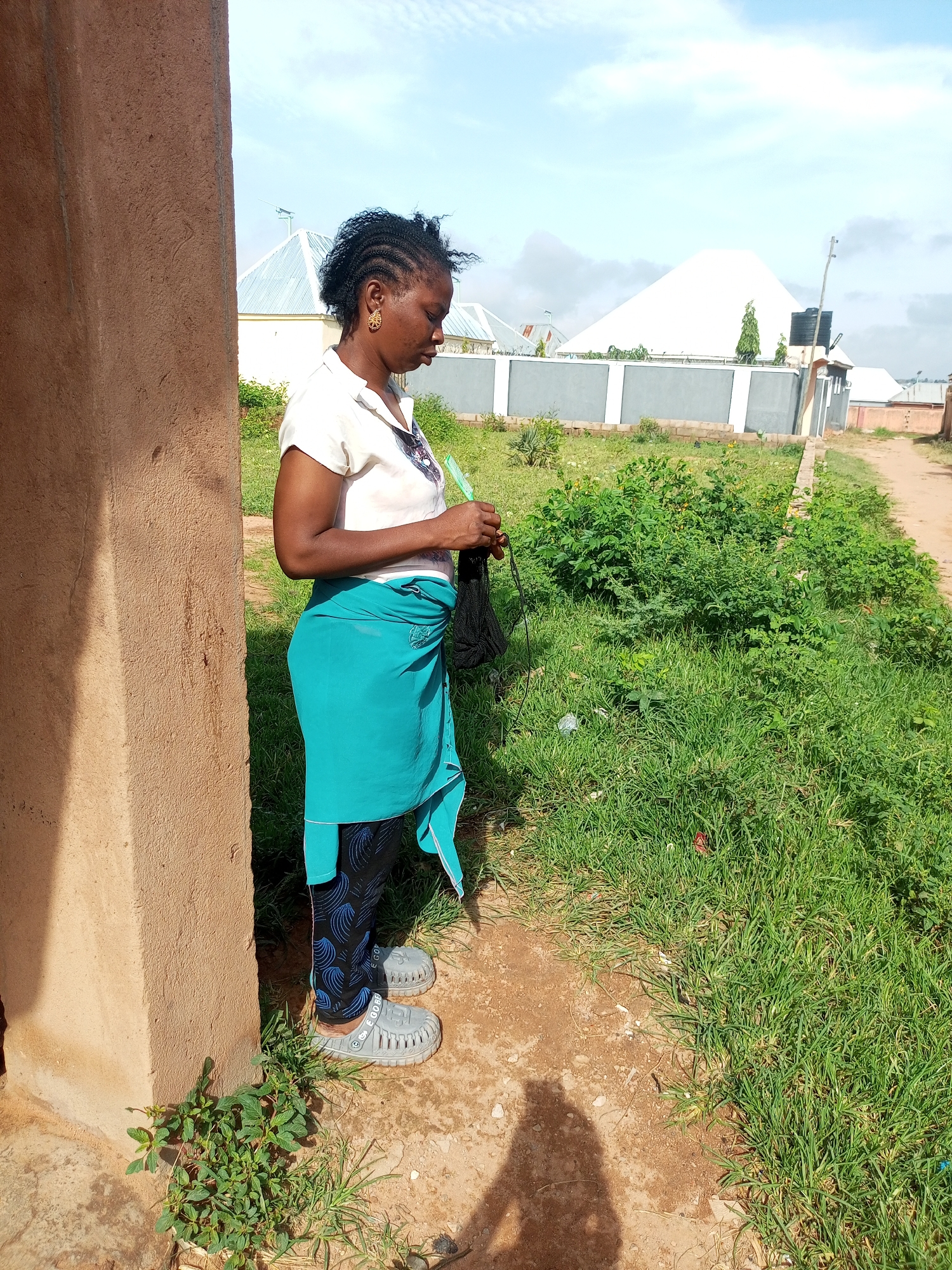 A full-shot, eye-level, outdoor medium shot captures a woman standing in profile, facing right, next to a textured, tan-colored wall. She wears a short-sleeved, white collared shirt, a turquoise wrap skirt tied around her waist, black leggings with a blue swirl pattern, and gray slip-on sandals. Her dark, braided hair is pulled back, and she has gold earrings. Her hands are engaged with a black object, possibly knitting or crocheting supplies. The background features a dirt path on the right, a lush green grassy area with scattered vegetation, and in the distance, a white, pyramid-shaped structure with a fence and some buildings. The sky is blue with scattered clouds.