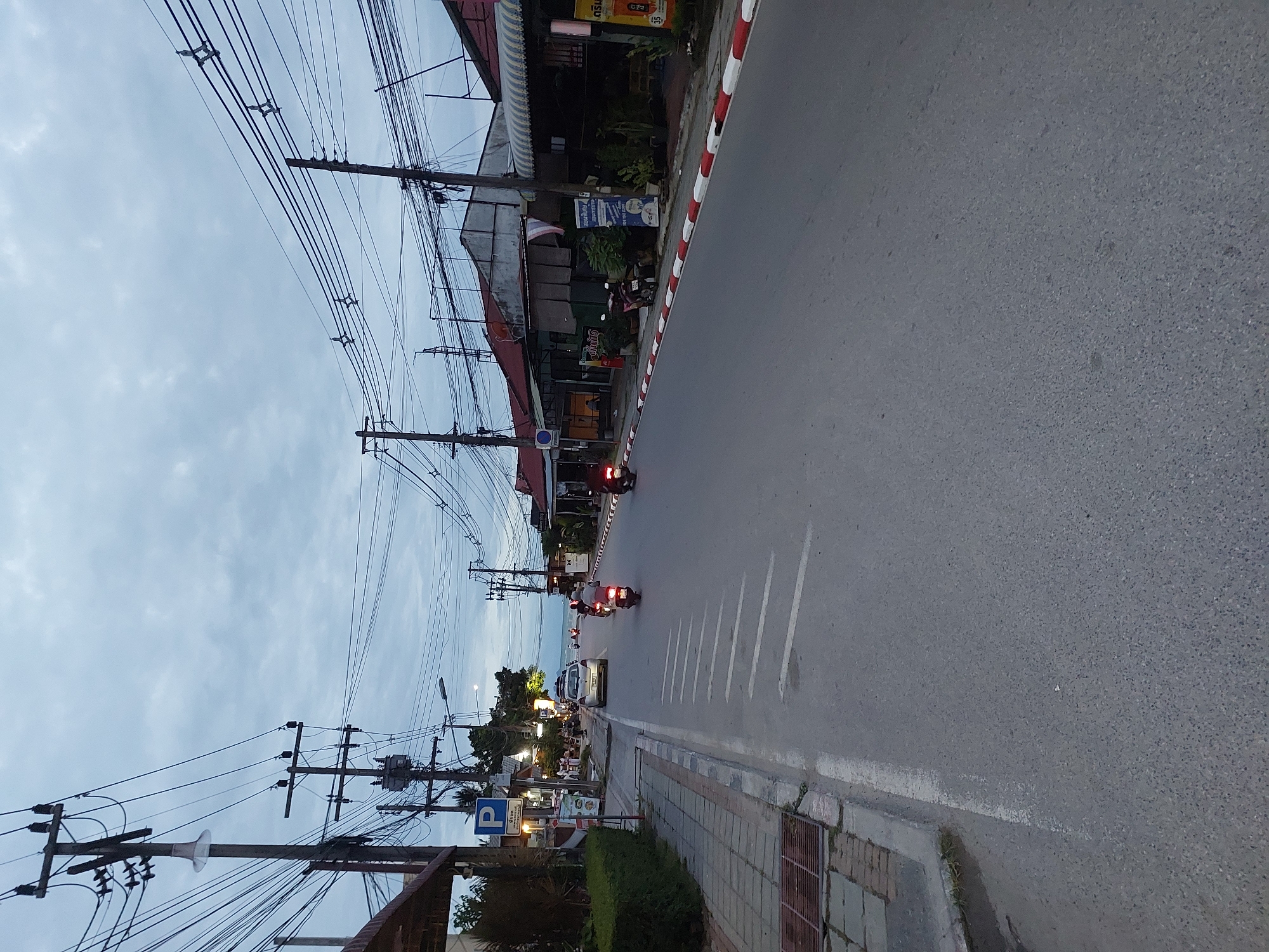 The image depicts a street scene in Ko Samui District, Thailand, during the evening or twilight hours. The sky is overcast with clouds. The street is paved with asphalt and marked with white lane lines, with a pedestrian sidewalk on the left. Several utility poles with a dense network of electrical wires are prominent along the street.
On the left side of the road, there are buildings with awnings and vegetation, including a small hedge. A blue parking sign is visible near a building. Several people are seated at tables outside one of the establishments.
On the road, a silver car is parked on the right side. Several motorcycles are present. Two motorcycles are actively being ridden by individuals wearing helmets. One motorcycle is to the right of the car, and another is further down the road on the right.
The buildings on the right side of the street appear to be shops or restaurants, with some signage and awnings. A red and white striped curb runs along the right side of the road. The overall impression is a developing commercial area with residential elements.
