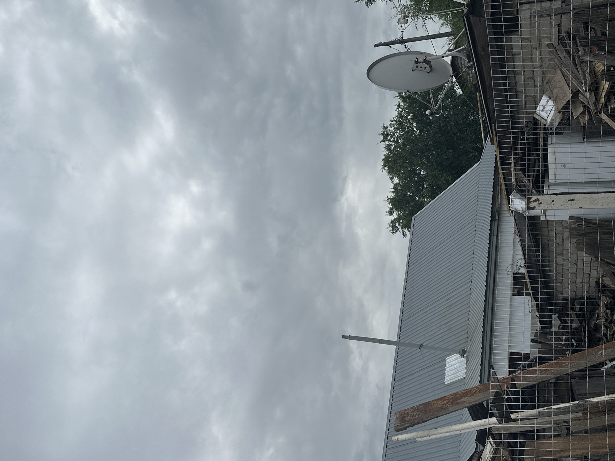The image depicts an outdoor scene under a cloudy sky. In the foreground, a wire mesh fence encloses a cluttered area with various construction debris, including wooden planks, bricks, and what appears to be a discarded refrigerator. Behind the fence, a low-rise building with a corrugated metal roof is visible, along with a metal chimney. To the right of the building, a satellite dish is mounted on a pole. Green trees form a backdrop behind the structures. The overall impression is one of a somewhat neglected or under-renovation area. The location is identified as the city of Tobyl, Kazakhstan. There are no people or animals visible in the image, and no specific actions or interactions are evident. The environment appears to be a residential or possibly a small industrial area.